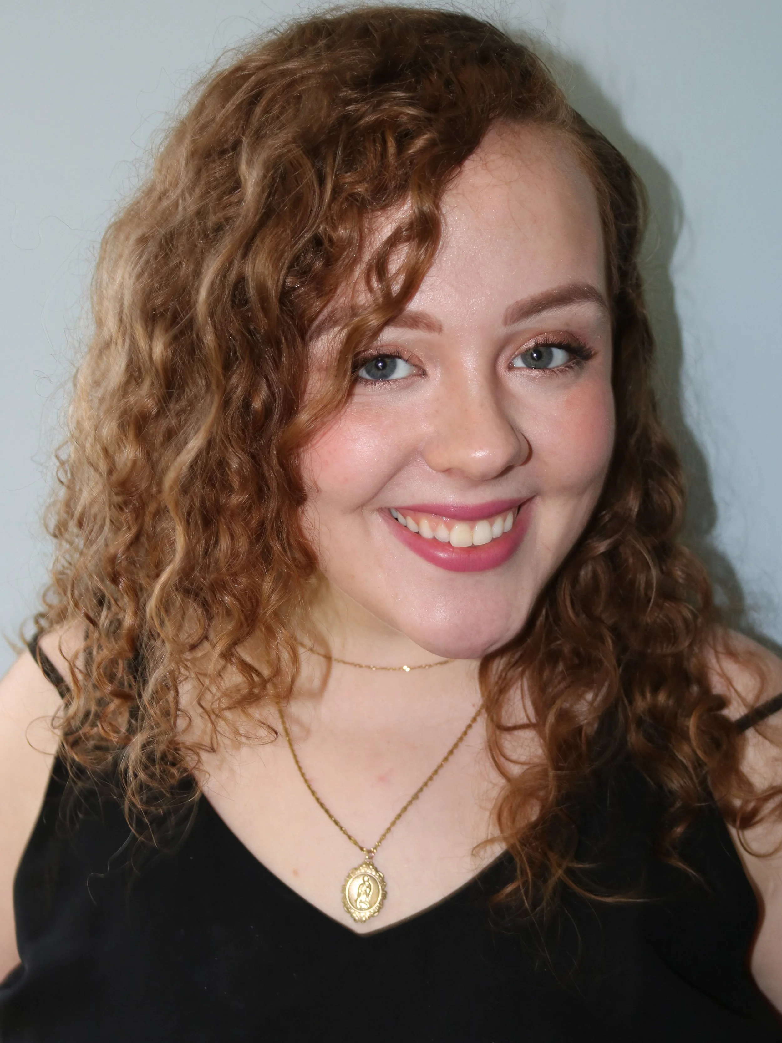 A young woman with curly red hair smiling, wearing a black top, a gold necklace with a religious pendant, and a thin gold choker. She has blue eyes and is standing against a plain, light-colored background.
