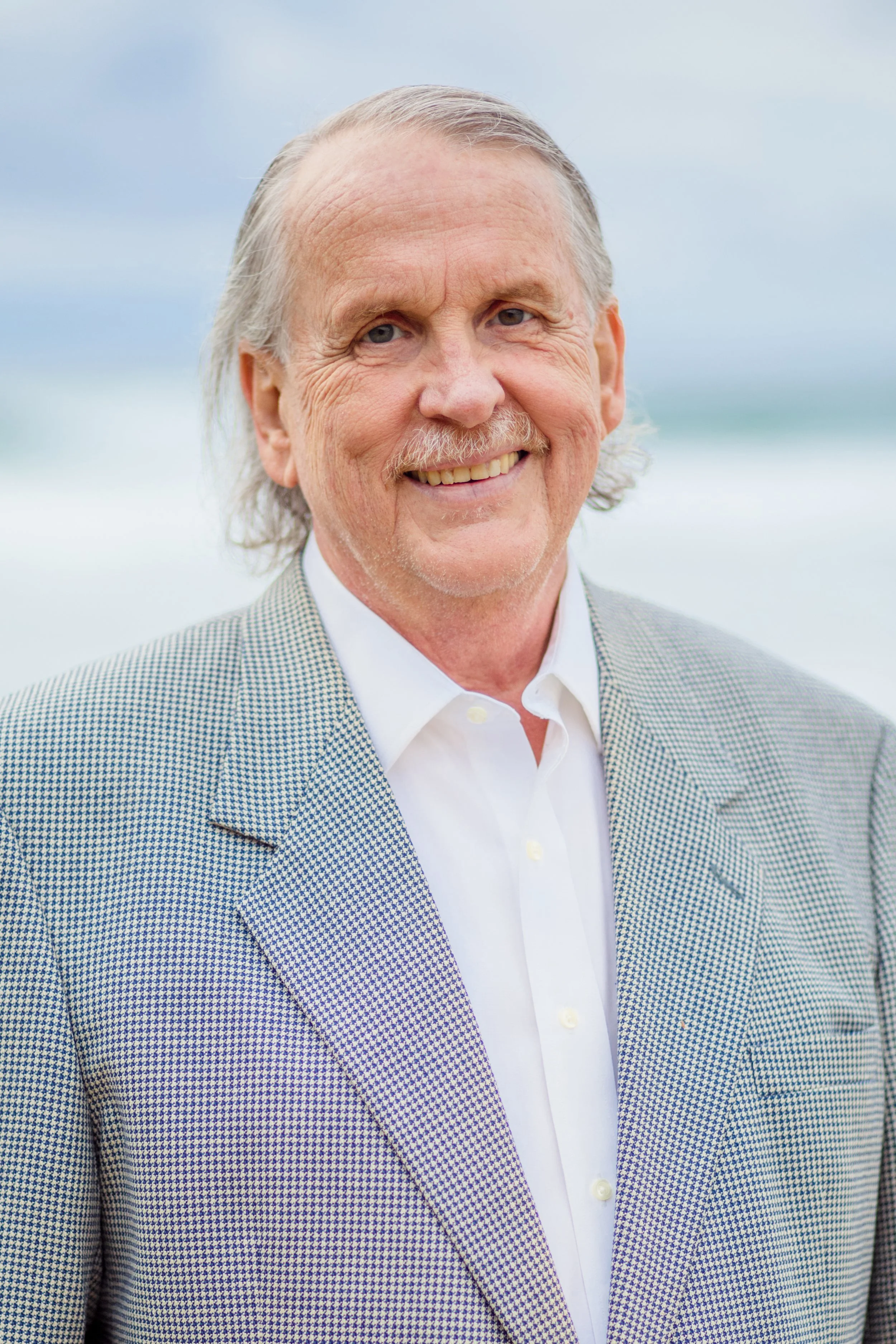 A smiling older man in a light-colored checkered blazer and white shirt, standing outdoors with a blurred beach and ocean background.