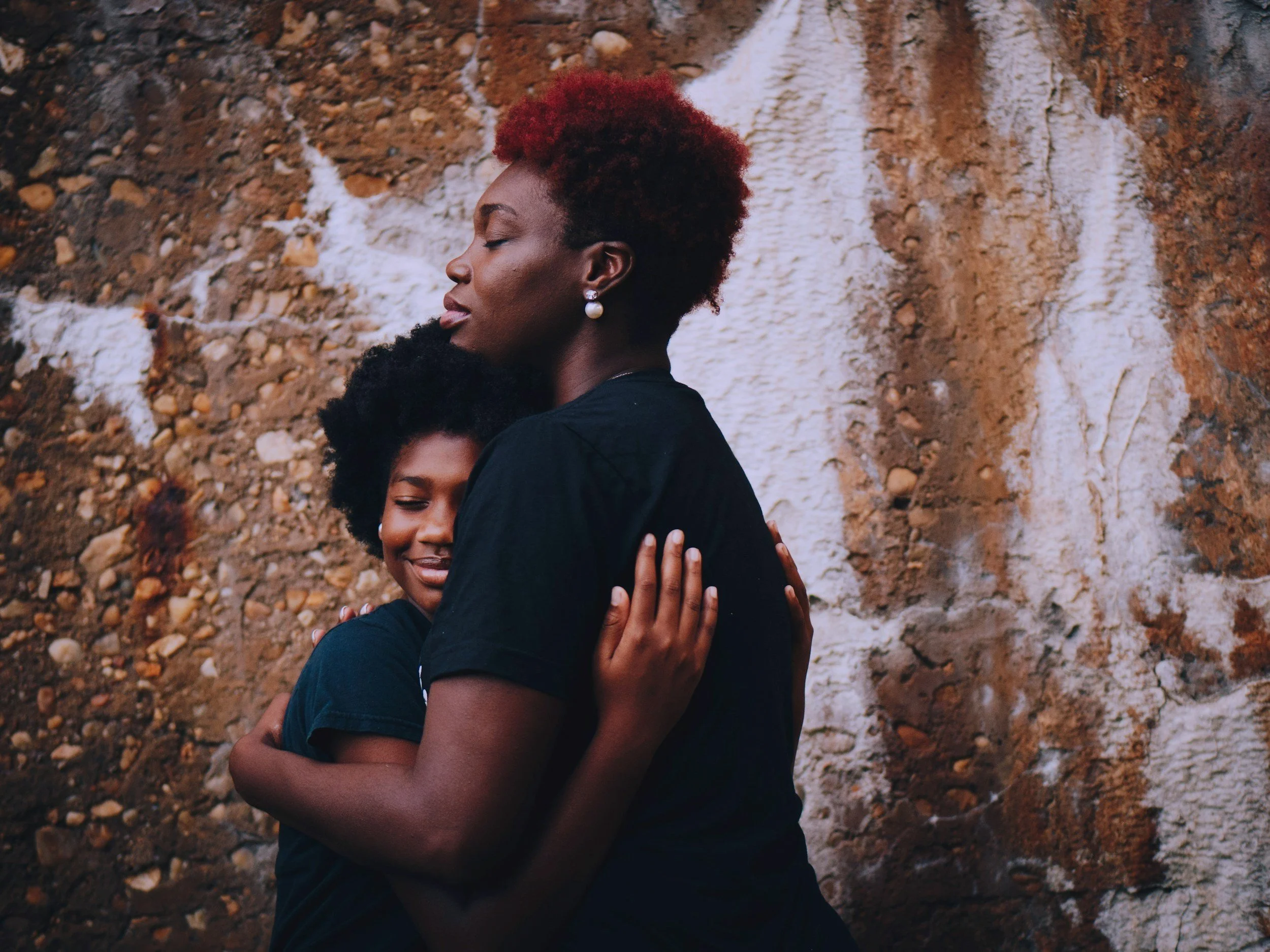 Two women hugging each other in front of a textured stone wall. The woman in front has reddish hair, closed eyes, and is wearing pearl earrings. The woman behind has curly black hair and a joyful expression.