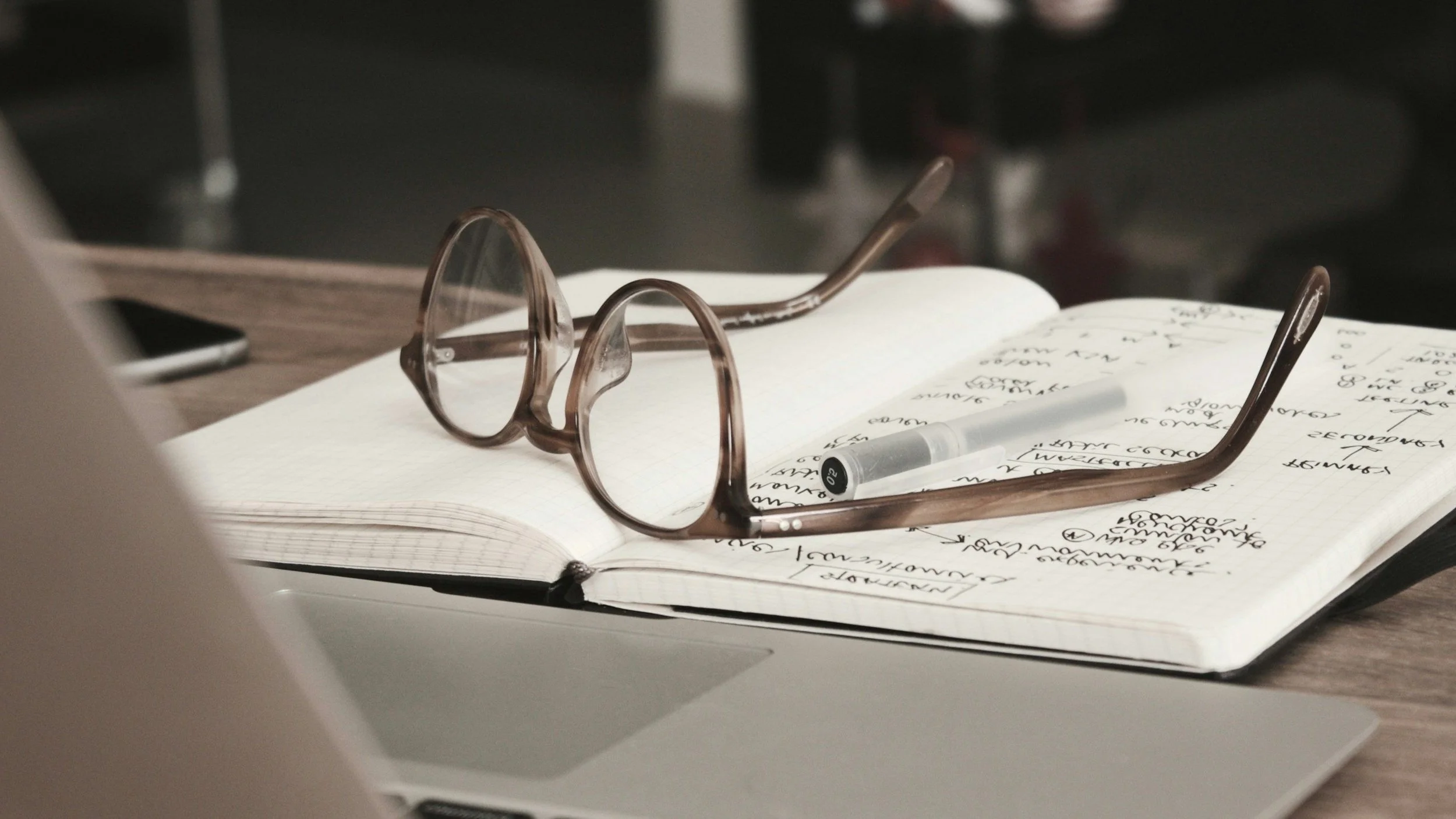 A pair of brown eyeglasses resting on an open notebook with handwritten notes and a white pen, on a wooden desk.