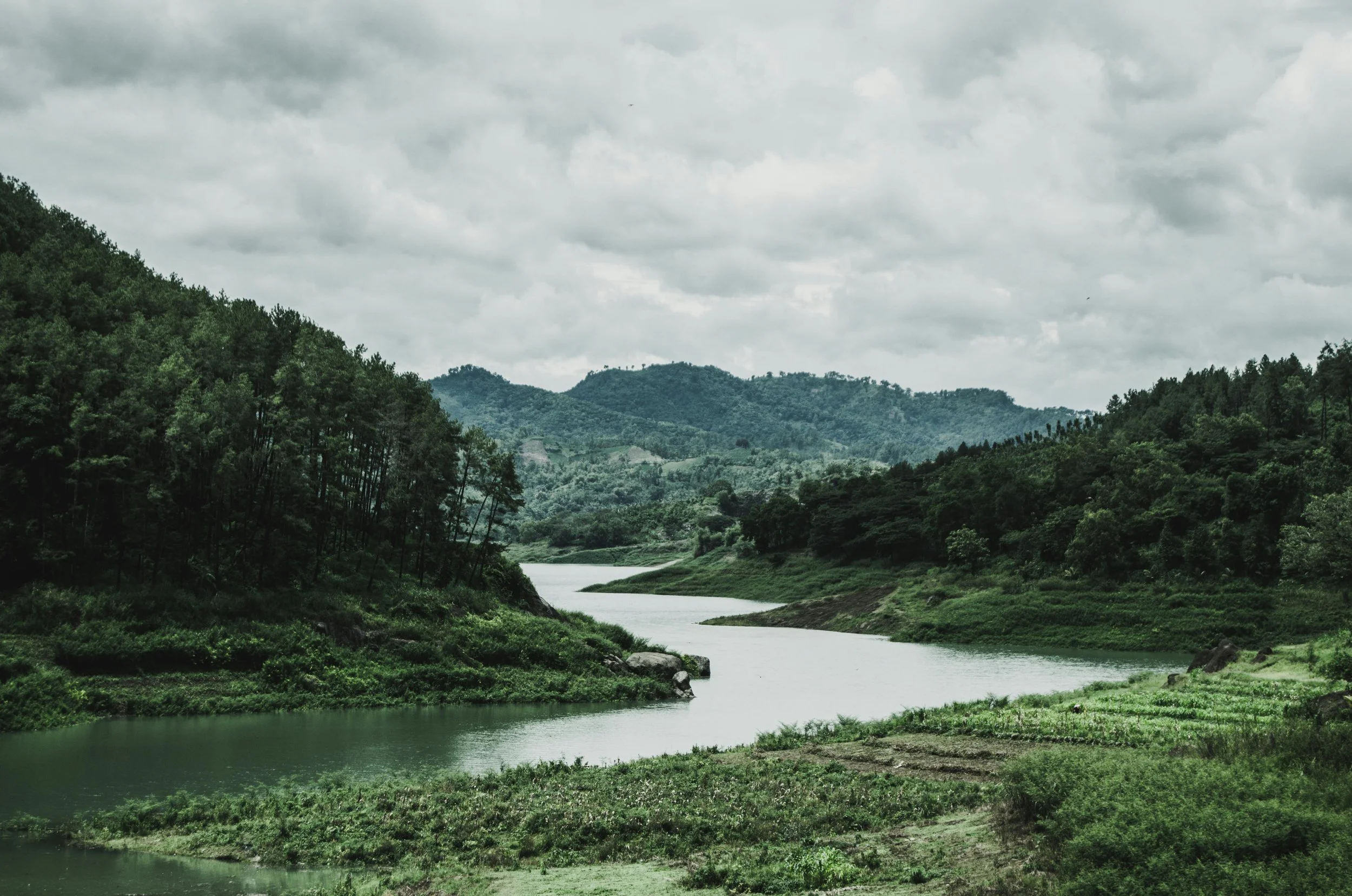 A scenic view of a winding river flowing through lush green hills and mountains under a cloudy sky.
