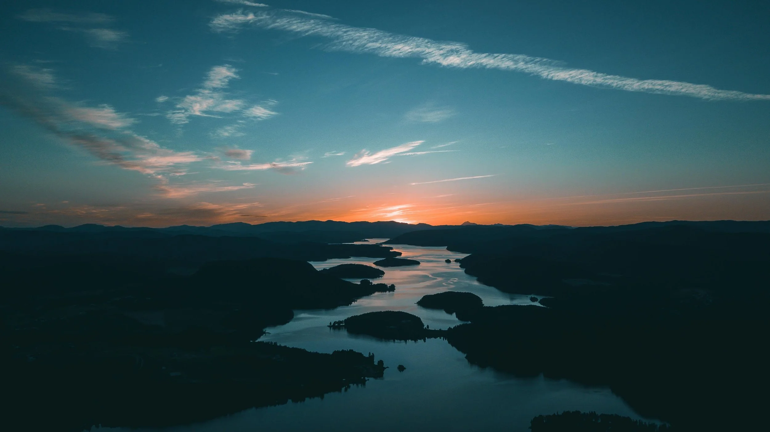Sunset over a meandering river flowing through a landscape of rolling hills and mountains, with a partly cloudy sky.