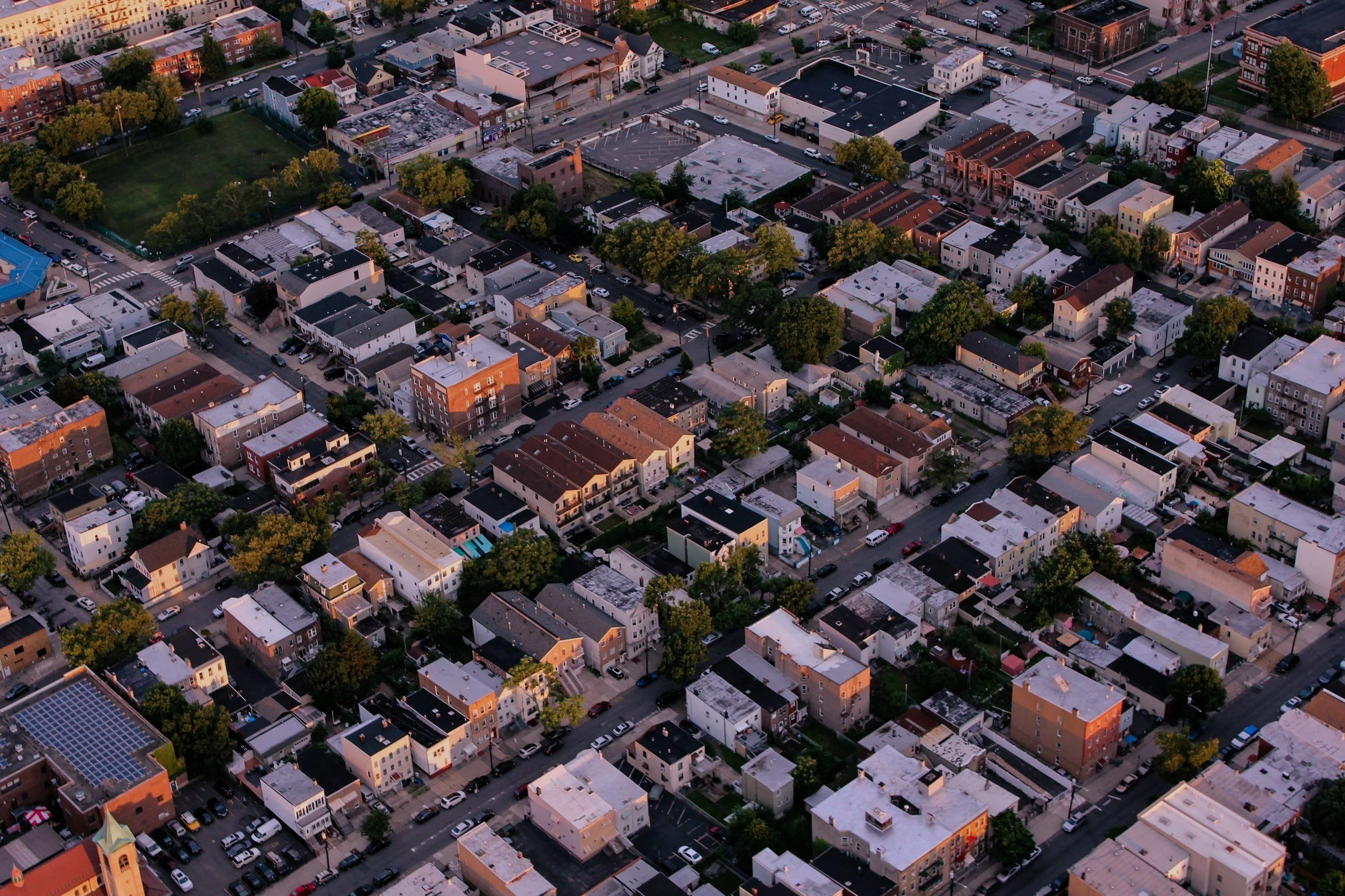 Aerial view of a densely packed urban neighborhood with a grid of streets, residential buildings, trees, and some parks during sunset or sunrise.