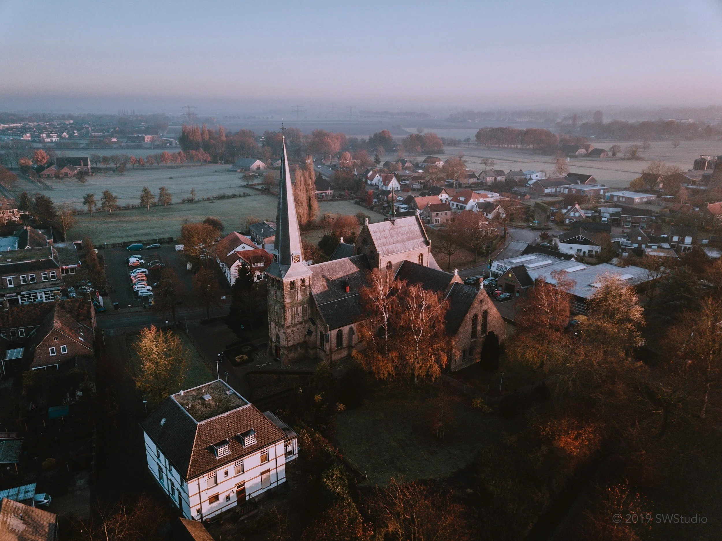 Aerial view of a small village with a church featuring a tall steeple, surrounded by houses and fields in a foggy, rural landscape during early morning or late afternoon.