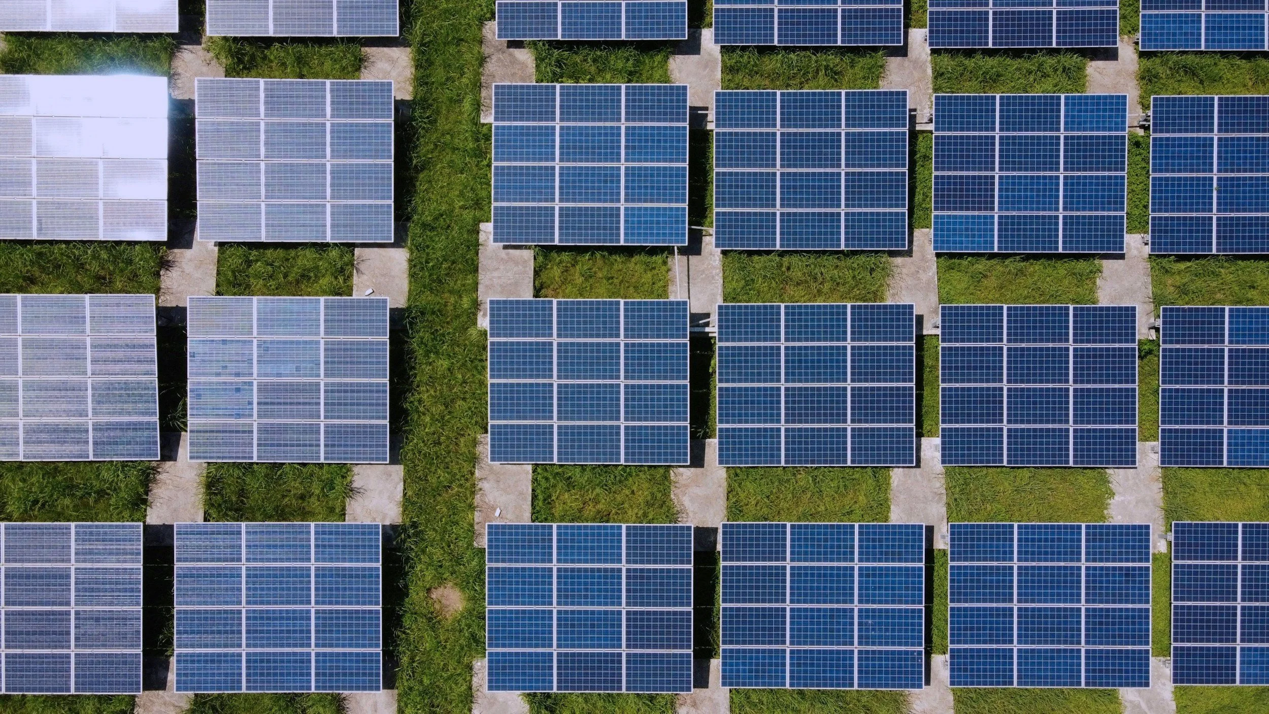 Aerial view of multiple solar panel arrays arranged in grid patterns with grass and gravel pathways between them.