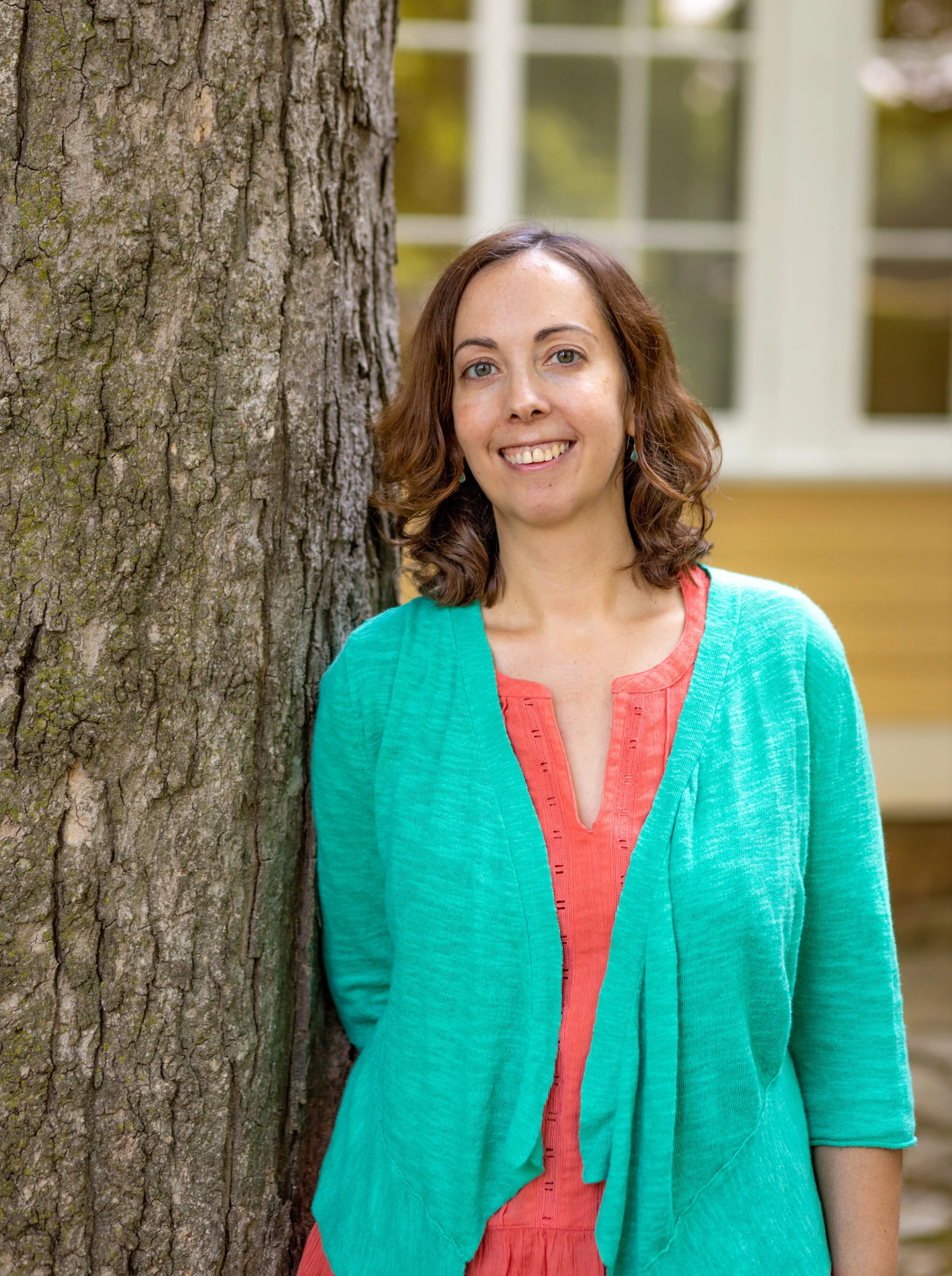 A woman with shoulder-length brown hair, wearing a turquoise cardigan over an orange top, standing outdoors beside a large tree trunk, smiling at the camera, with a house and windows in the background.