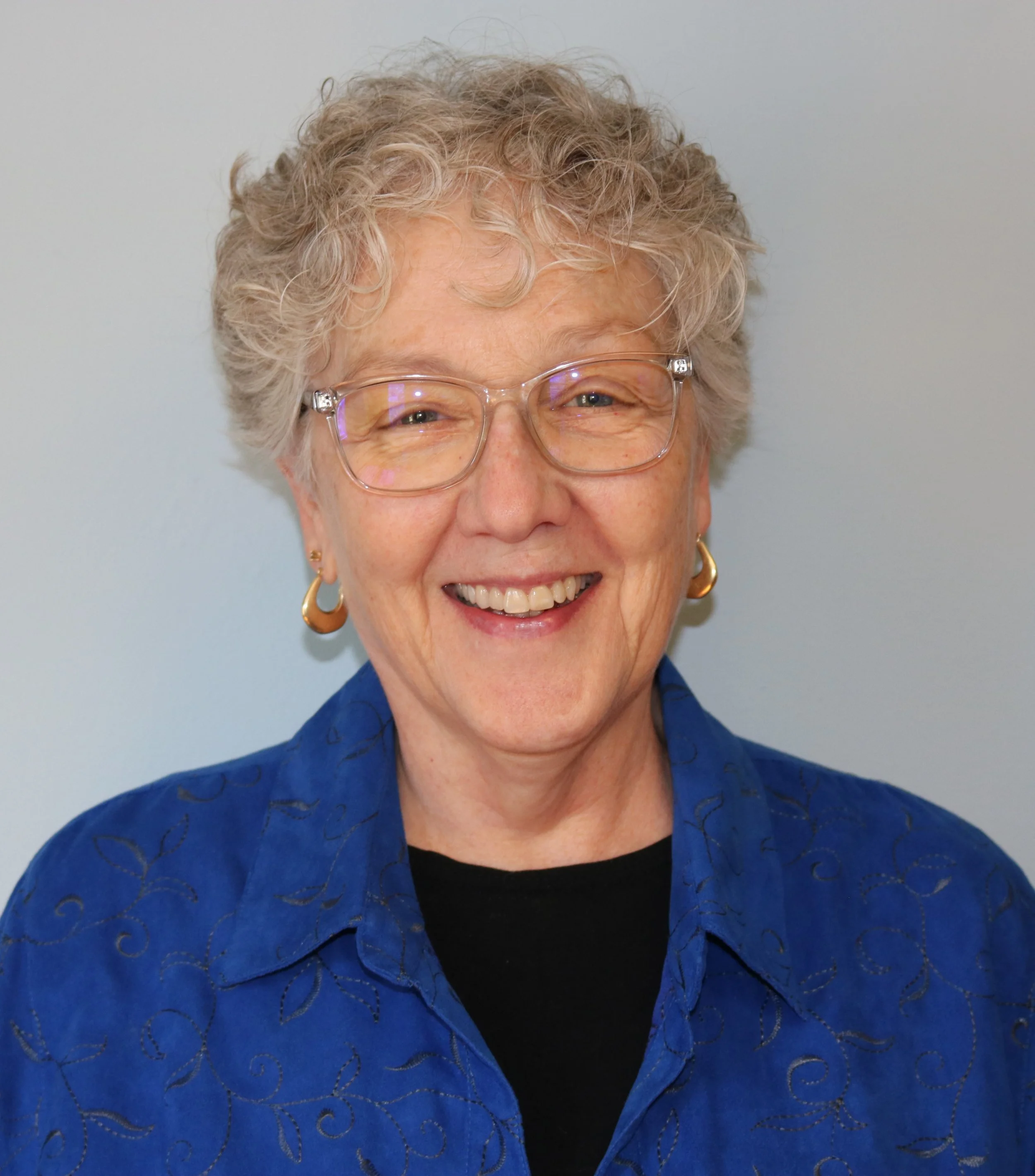 An older woman with curly gray hair and glasses, smiling, wearing a blue embroidered shirt and gold earrings.