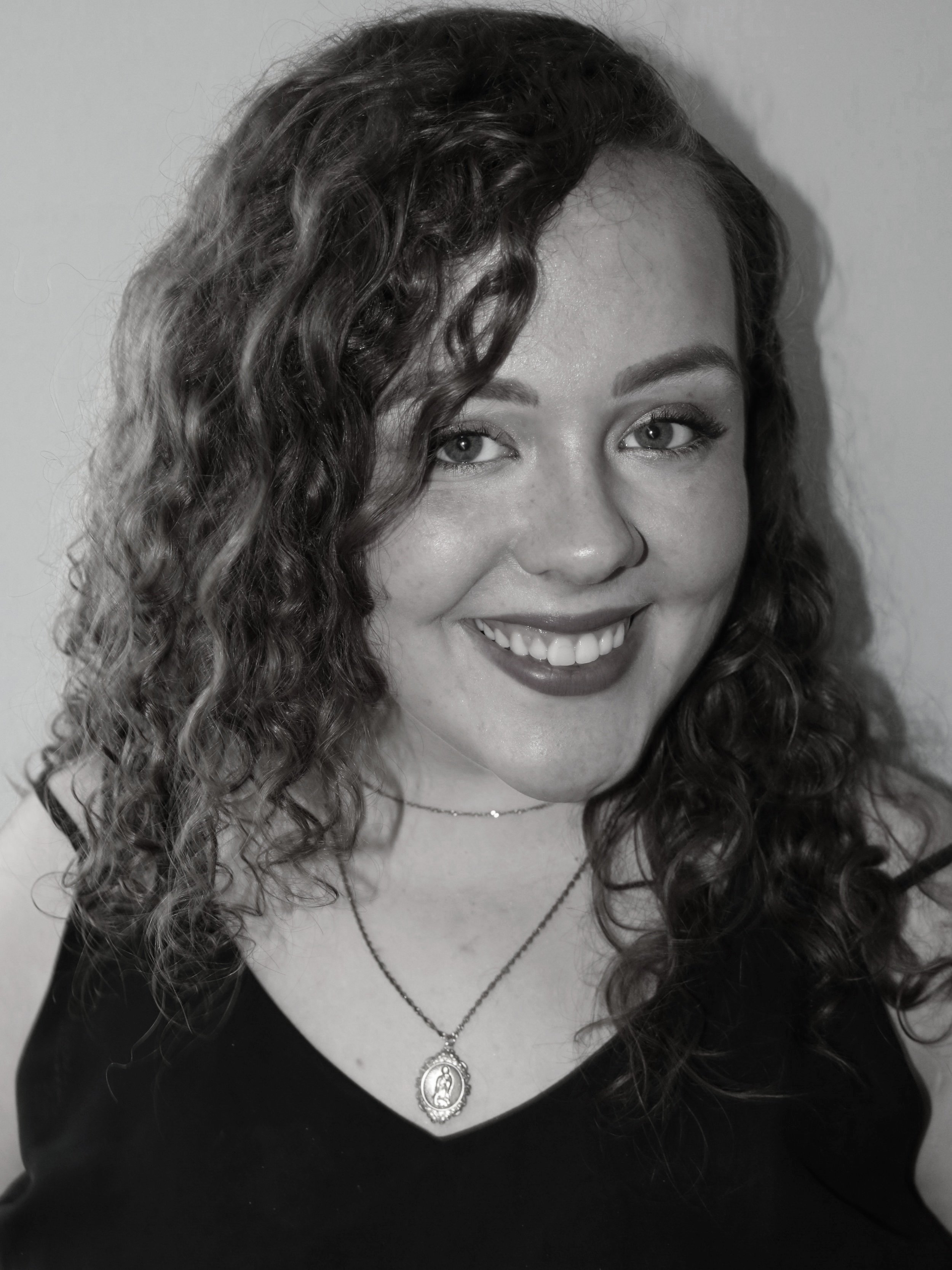 A woman with curly hair smiling in a black top, wearing a necklace with a pendant, photographed against a plain background.