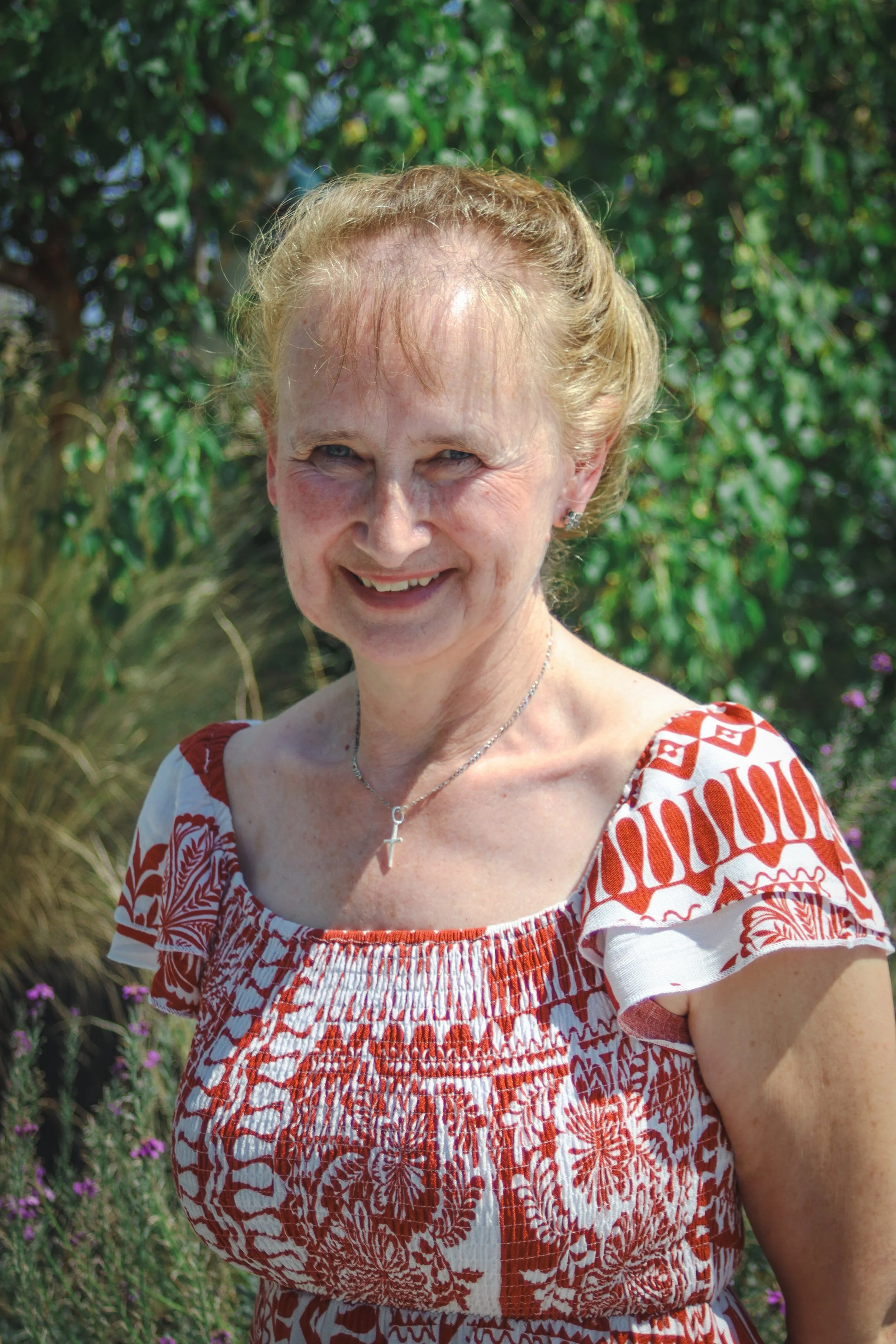 A woman with blonde hair in a bun, wearing a purple blazer and a multi-colored patterned blouse, smiling against a white background.