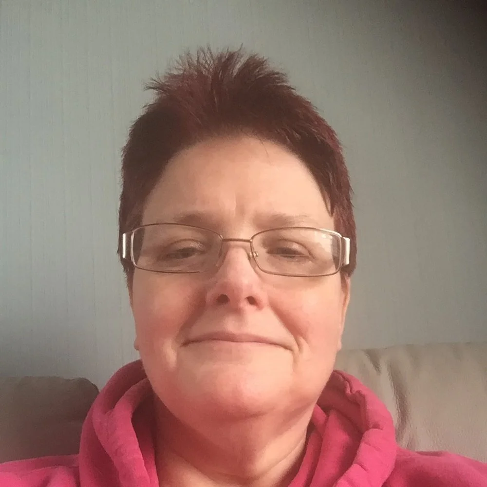 A person with short hair and glasses wearing a pink hoodie, sitting on a sofa.