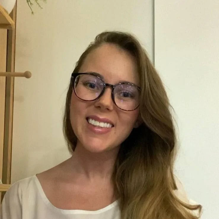 A woman with long brown hair and glasses smiling, wearing a white top, in a room with light-colored walls.