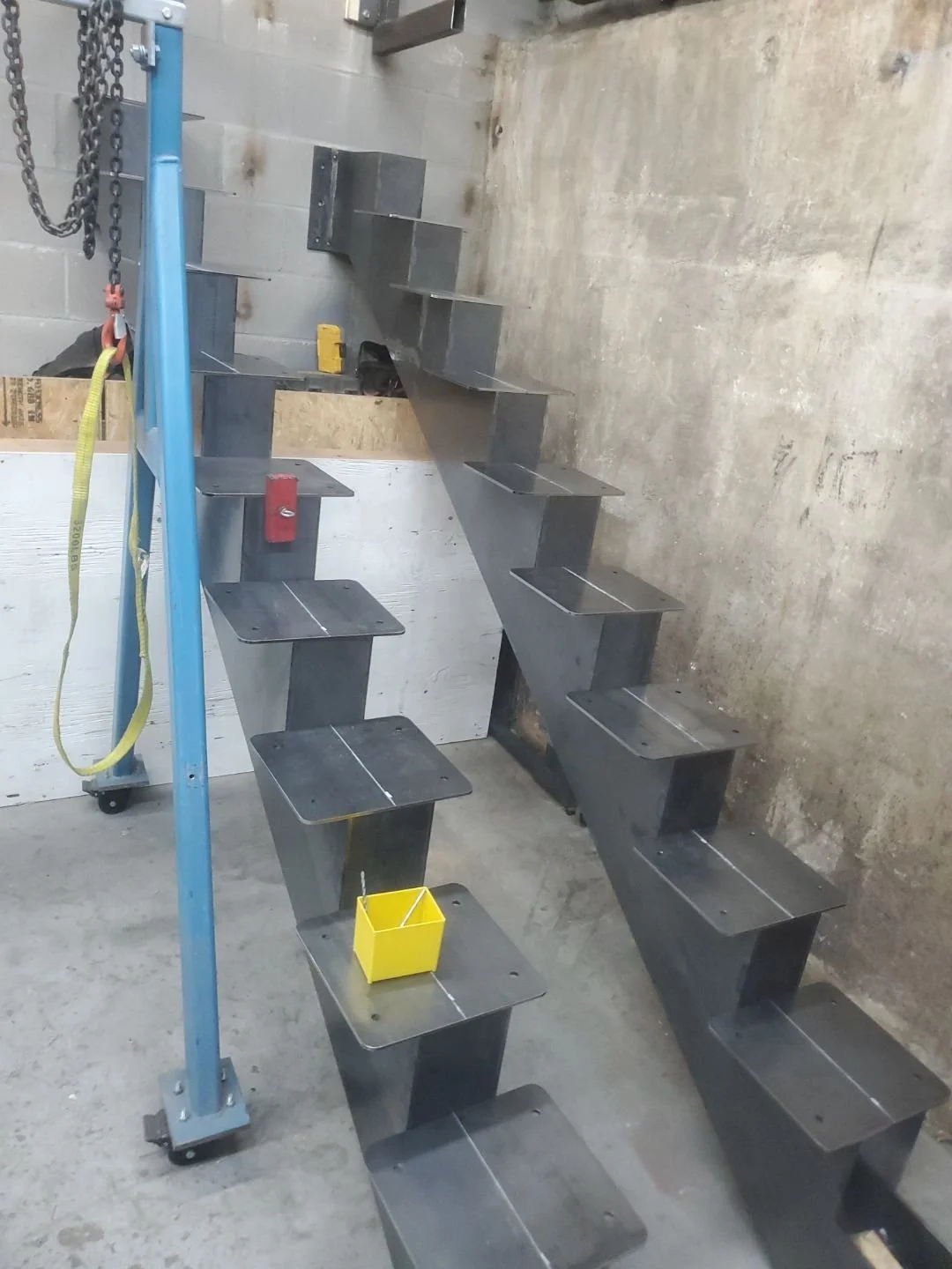 Metal staircase with alternating steps in an industrial setting, blue support beam, yellow container on step.
