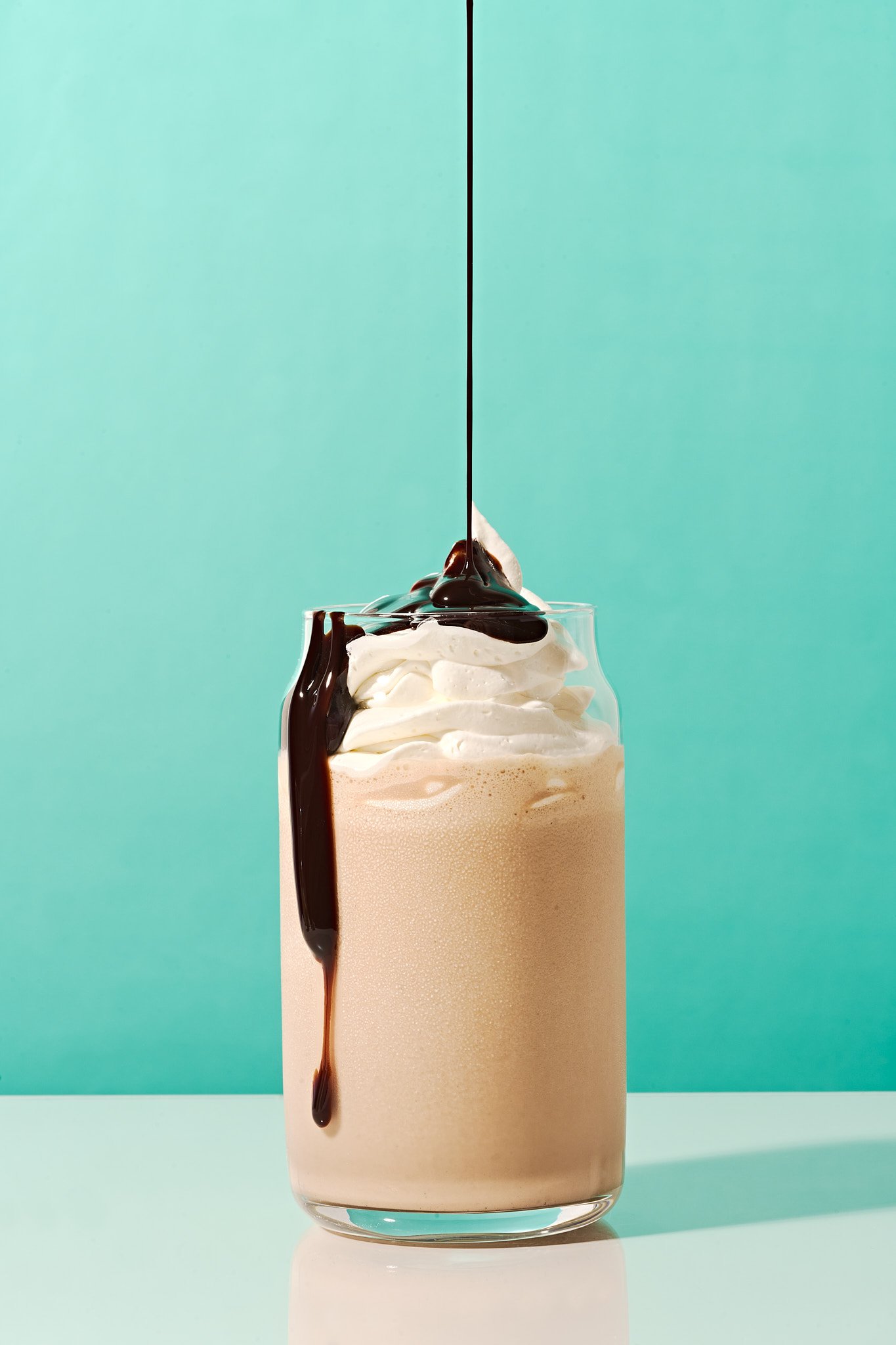 A glass of cold coffee topped with whipped cream and drizzled with chocolate syrup, with chocolate syrup being poured over the top. commercial food photography product photographer freelance consumer brand 