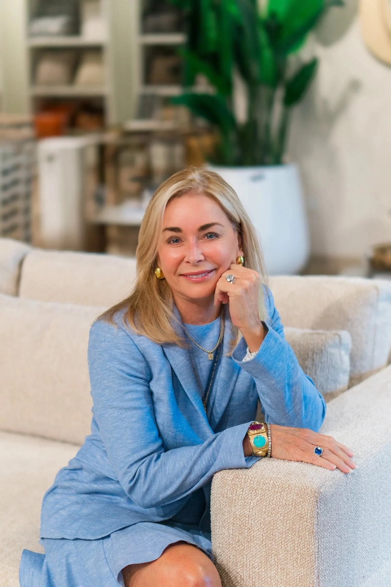 A woman with blonde hair and blue eyes sitting on a cream-colored sofa in a cozy room with a large green plant in the background. She is wearing a light blue blazer and has various jewelry, including rings, earrings, a necklace, and a bracelet. She is smiling and resting her chin on her hand.
