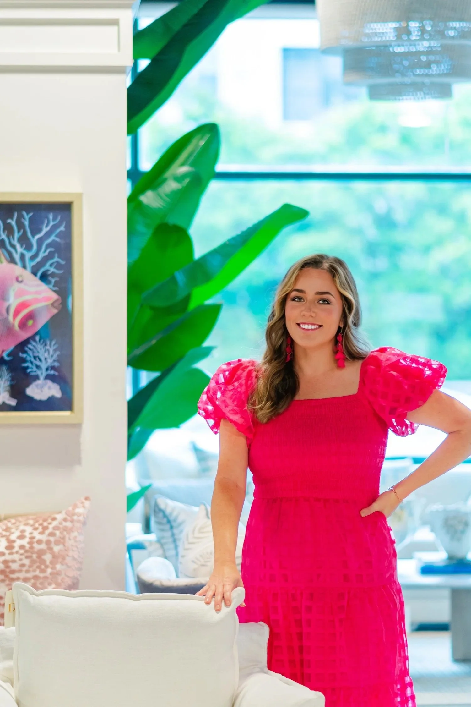 A woman with wavy brown hair, smiling, wearing a bright pink dress with puffed sleeves and pink earrings, standing in a modern, well-lit indoor setting with large green plants and artwork in the background.