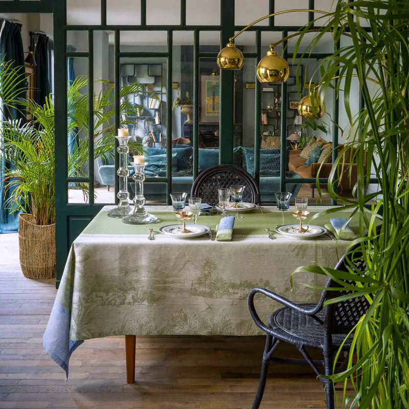 A dining room with a table set for a meal, surrounded by black chairs, with large potted plants, and a glass divider separating it from a living room featuring a blue sofa and various decor.