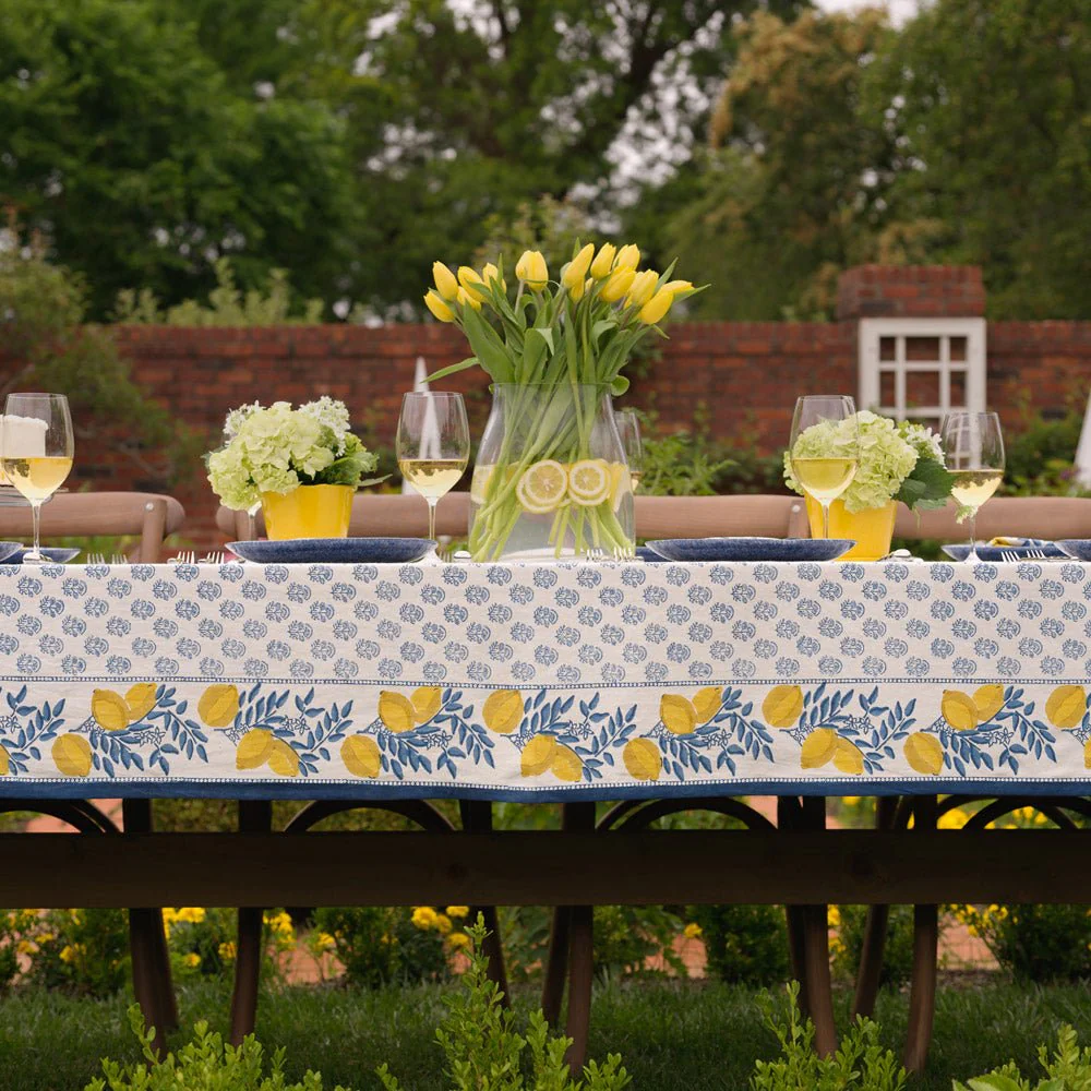 Outdoor dining table decorated with a lemon-themed tablecloth, yellow tulips in a glass vase, and glasses of white wine, set against a garden background with a red brick wall and greenery.