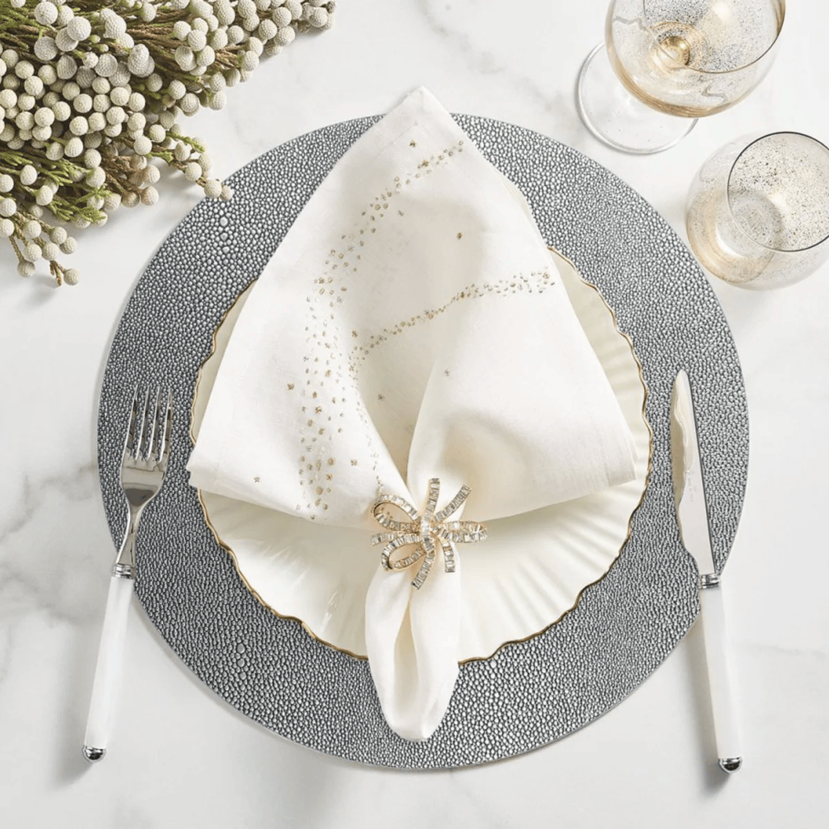 Elegant table setting with a silver patterned charger plate, white napkin folded on top, decorated with a jewel brooch, silver fork and knife on white handles, two glasses of white wine or champagne, and a bouquet of white flowers in the upper left corner.