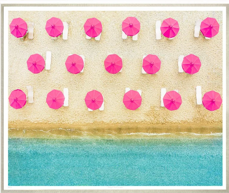 Beach with pink umbrellas and white lounge chairs, viewed from above.