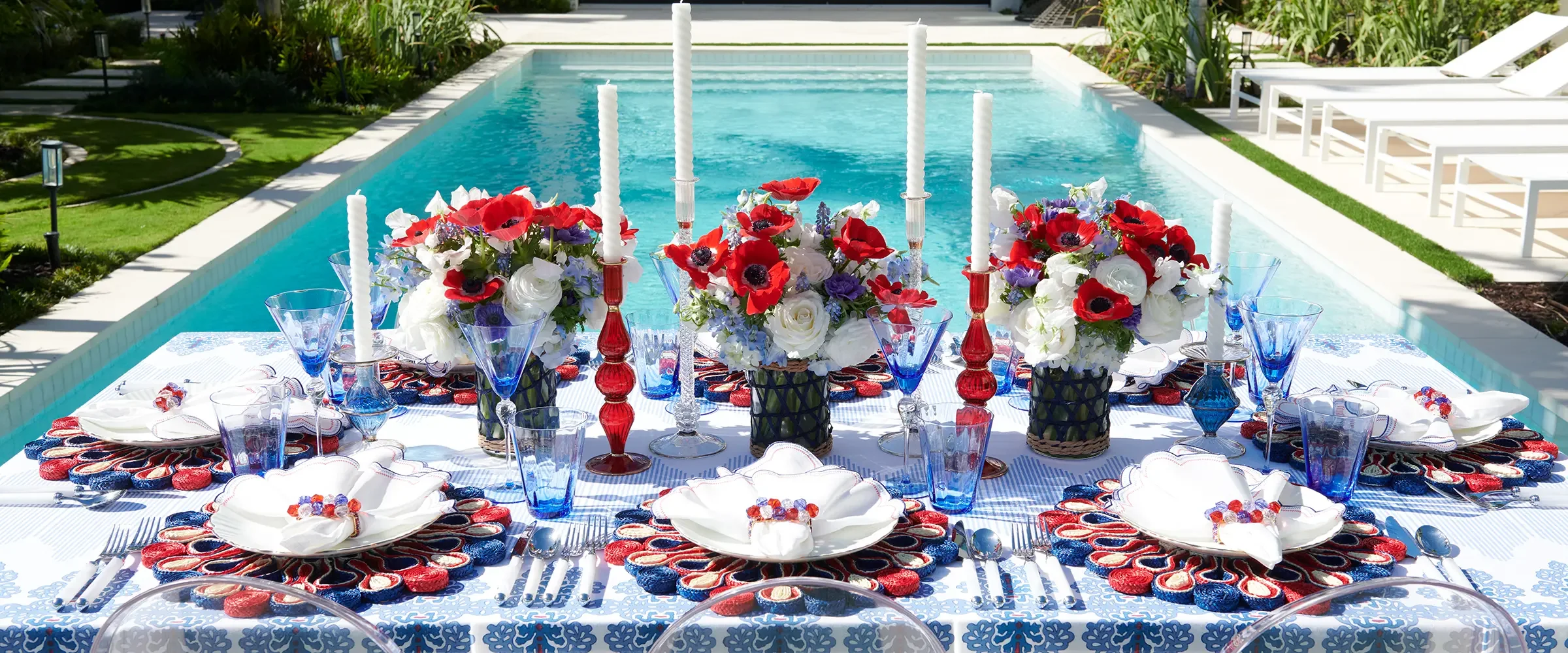 A decorated outdoor table set with flowers, candles, glassware, and plates, in front of a swimming pool with lounge chairs.