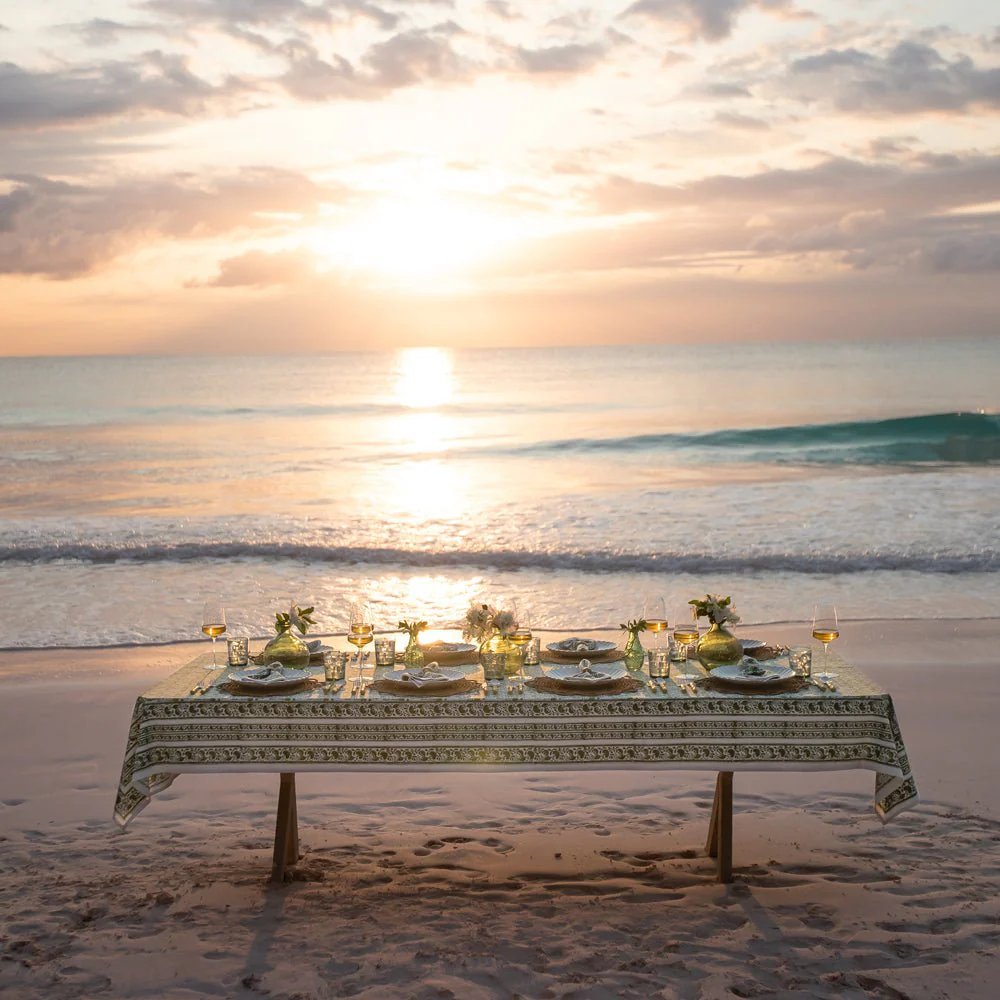 A table set for a meal on a beach at sunset, with the ocean and sky in the background.