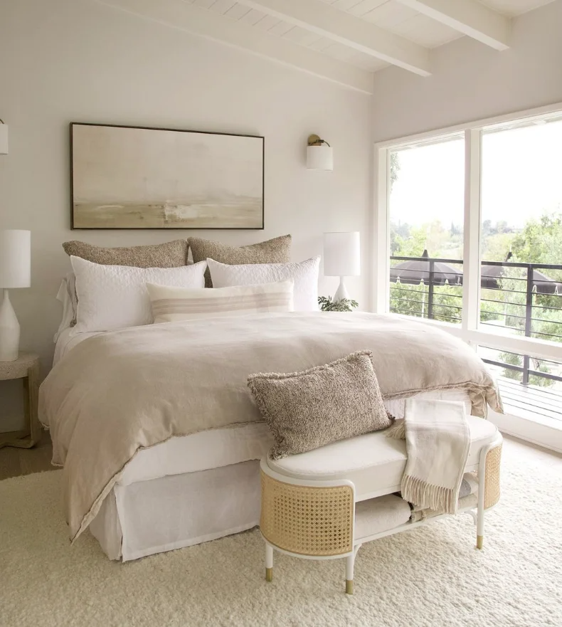 Bright bedroom with a large bed covered in beige and white linens, decorative pillows, and a bench at the foot of the bed. Two nightstands with white lamps flank the bed. A large sliding glass door leads to a balcony with a view of trees and outdoor