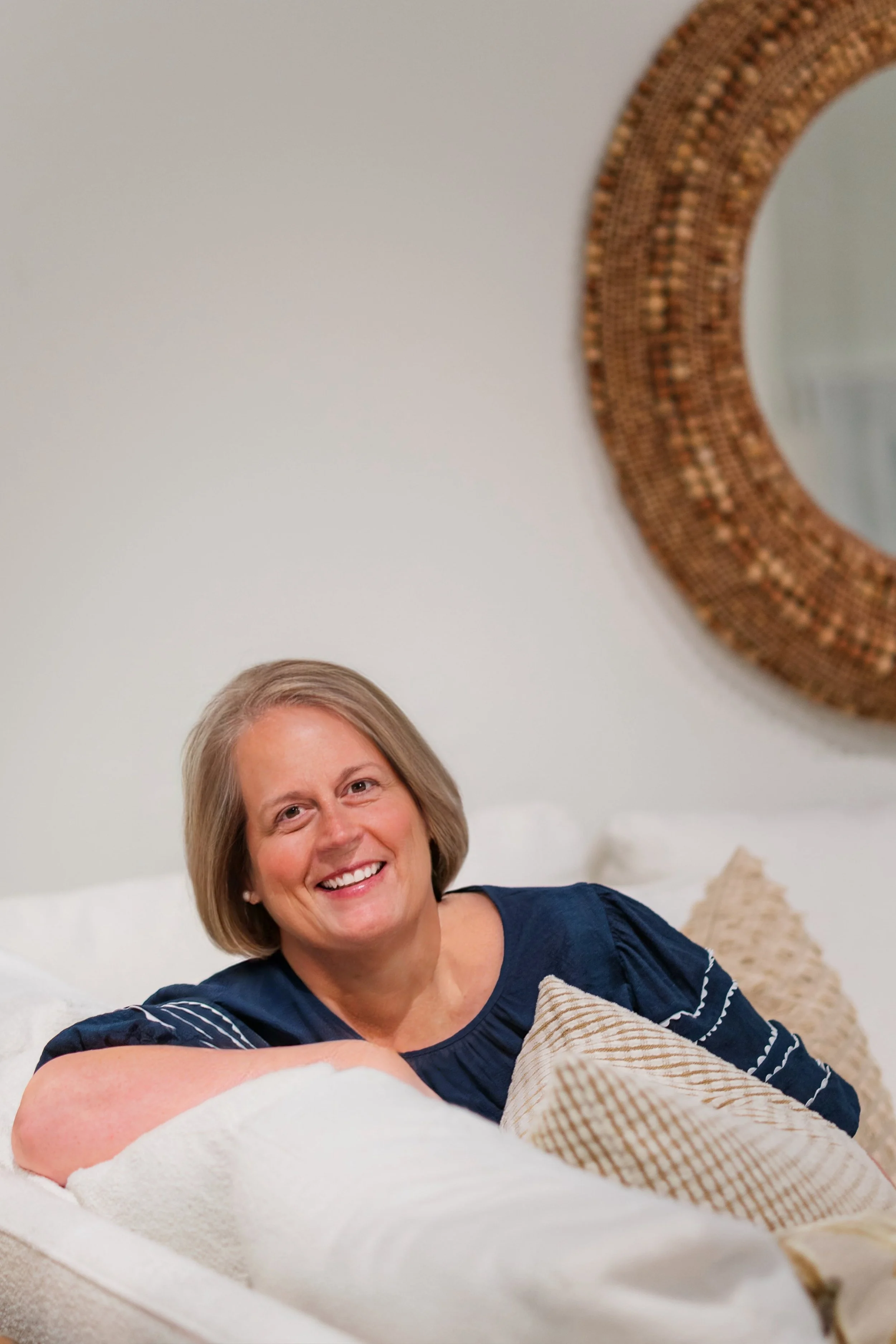 A middle-aged woman with short blonde hair smiling, sitting on a white couch with decorative pillows, in a room with a large round mosaic mirror on the wall behind her.