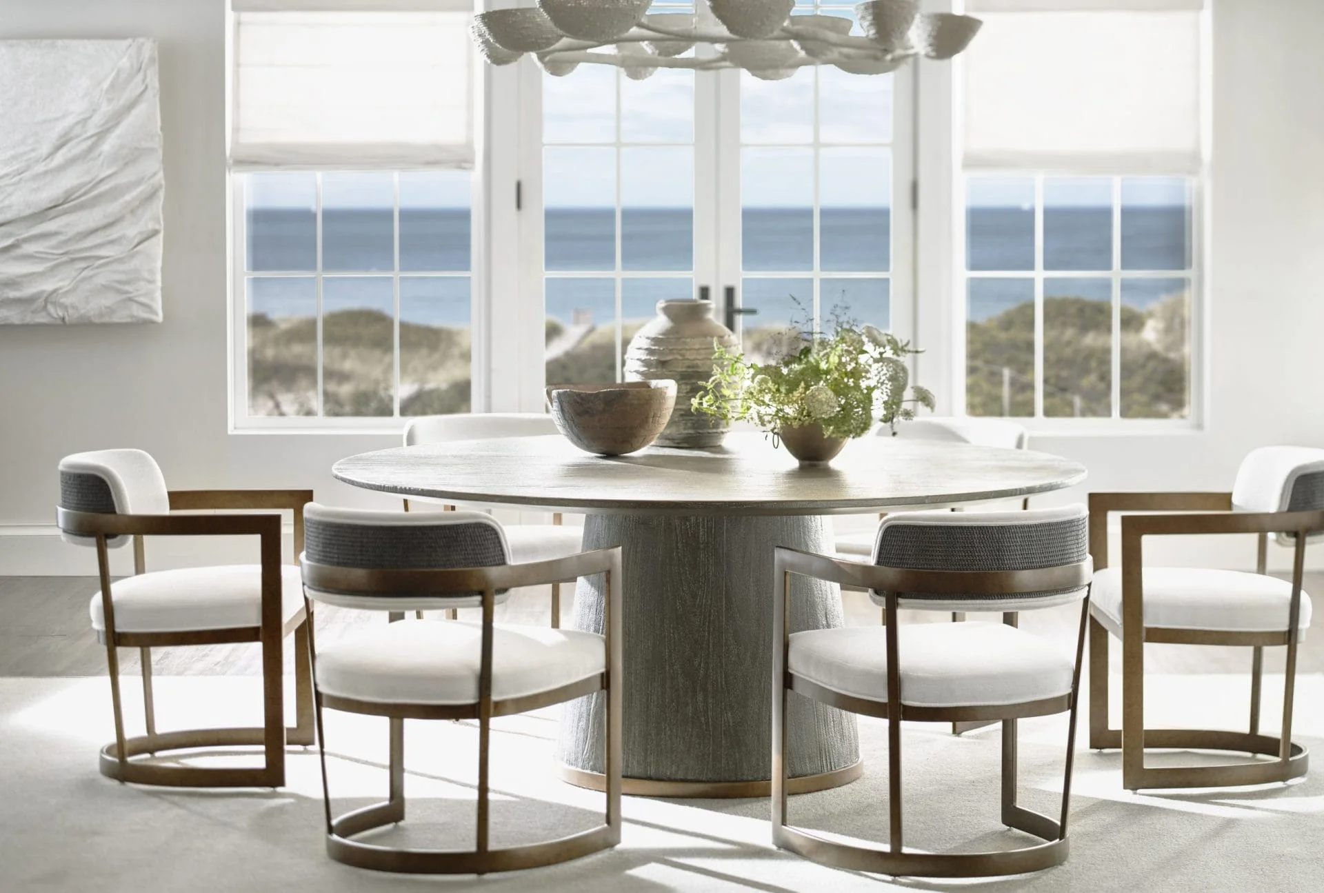 A bright dining room with a round wooden table, six chairs with white cushions, a view of the ocean through large windows, and decorative vases and a plant on the table.