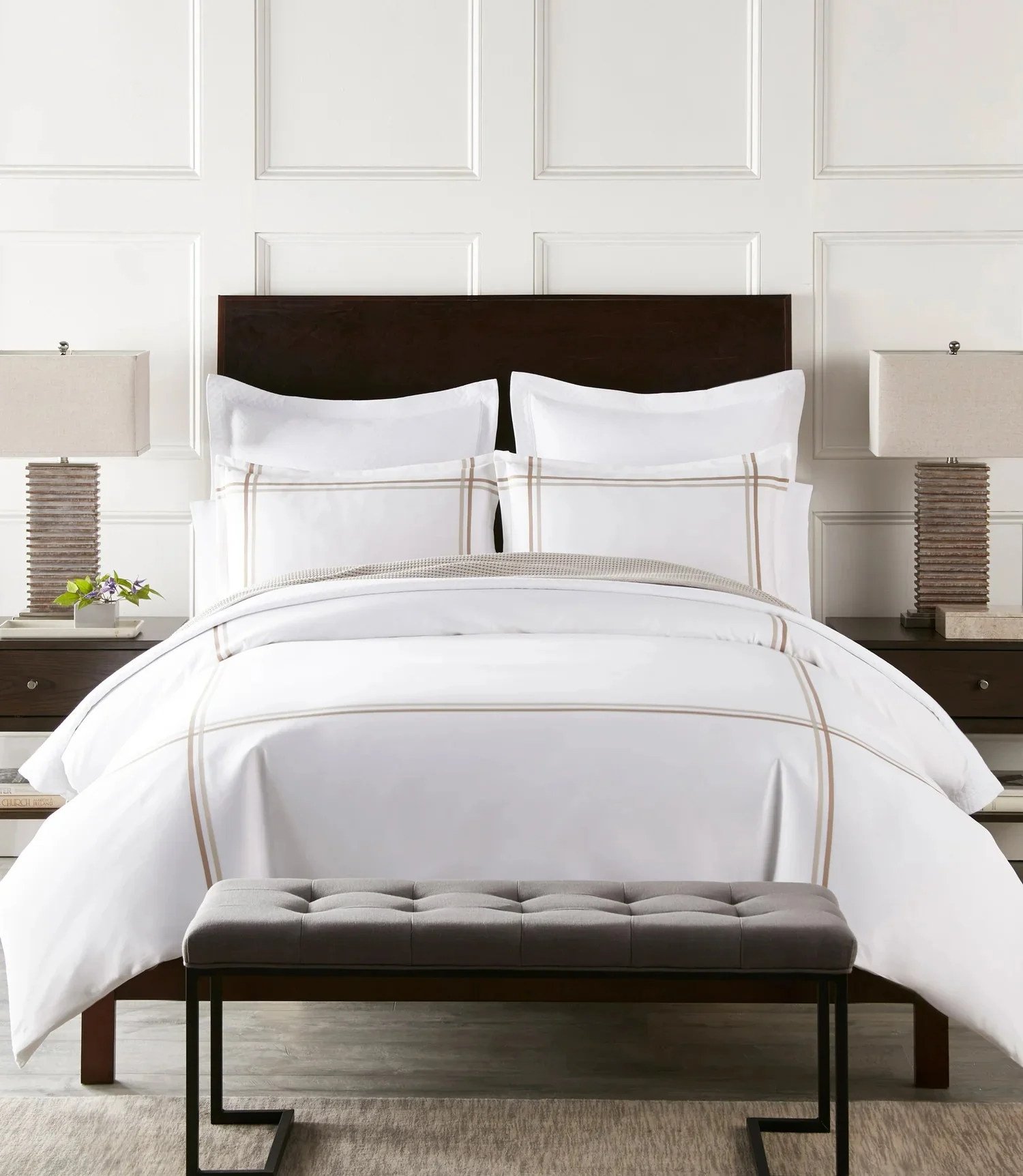 A neatly made bed with white bedding and beige checkered accents, flanked by two bedside tables with matching lamps, a small plant on the left nightstand, and a gray upholstered bench at the foot of the bed, all in a modern bedroom.