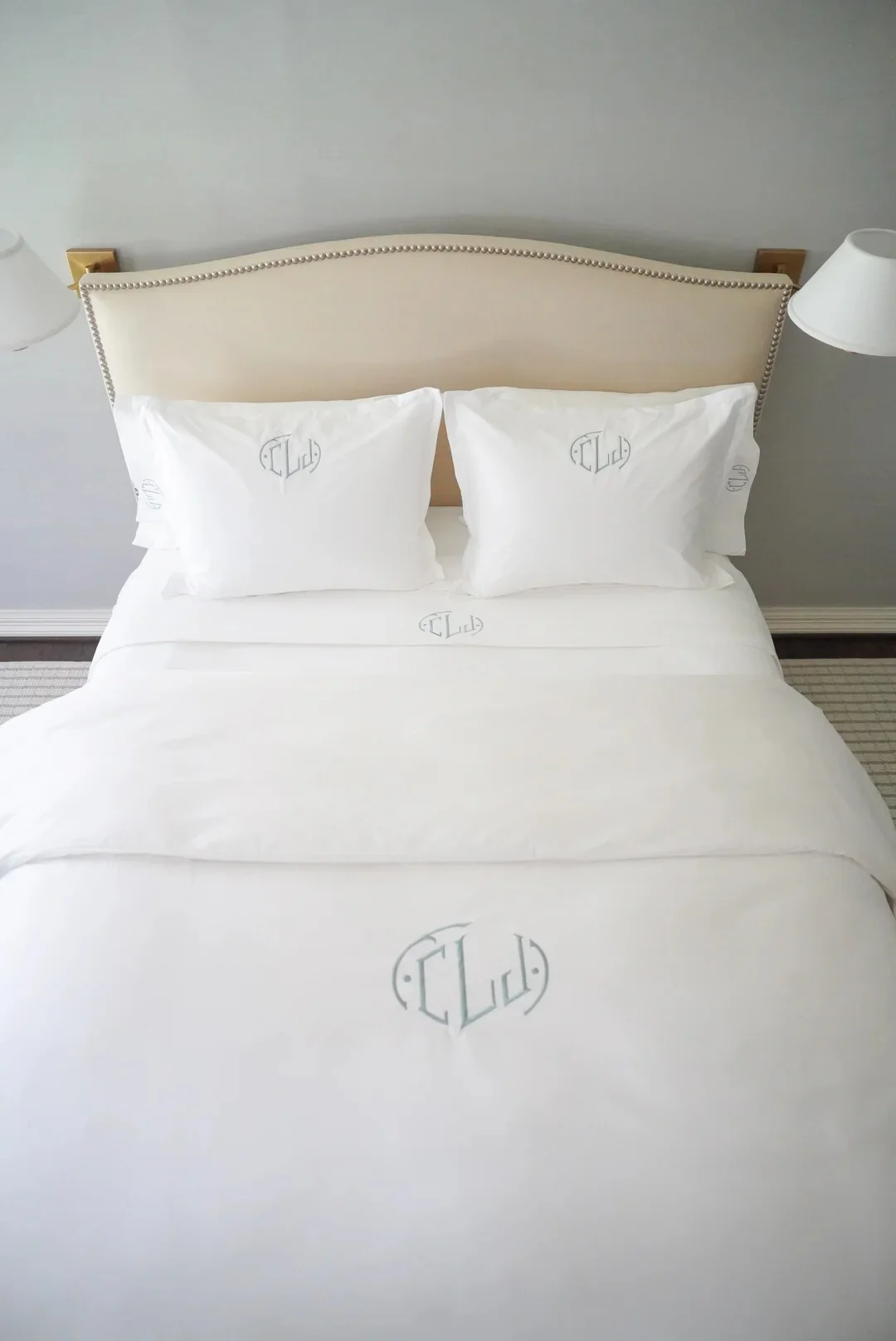 A neatly made bed with white sheets, two pillows with embroidered logos, beige upholstered headboard, and wall-mounted lamps on both sides.