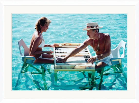 Two elderly people playing checkers on a floating table in a swimming pool.