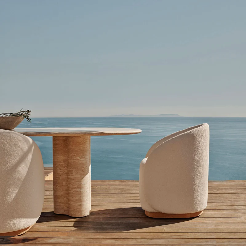 A wooden outdoor deck overlooking the ocean with a white upholstered chair and a round stone table.