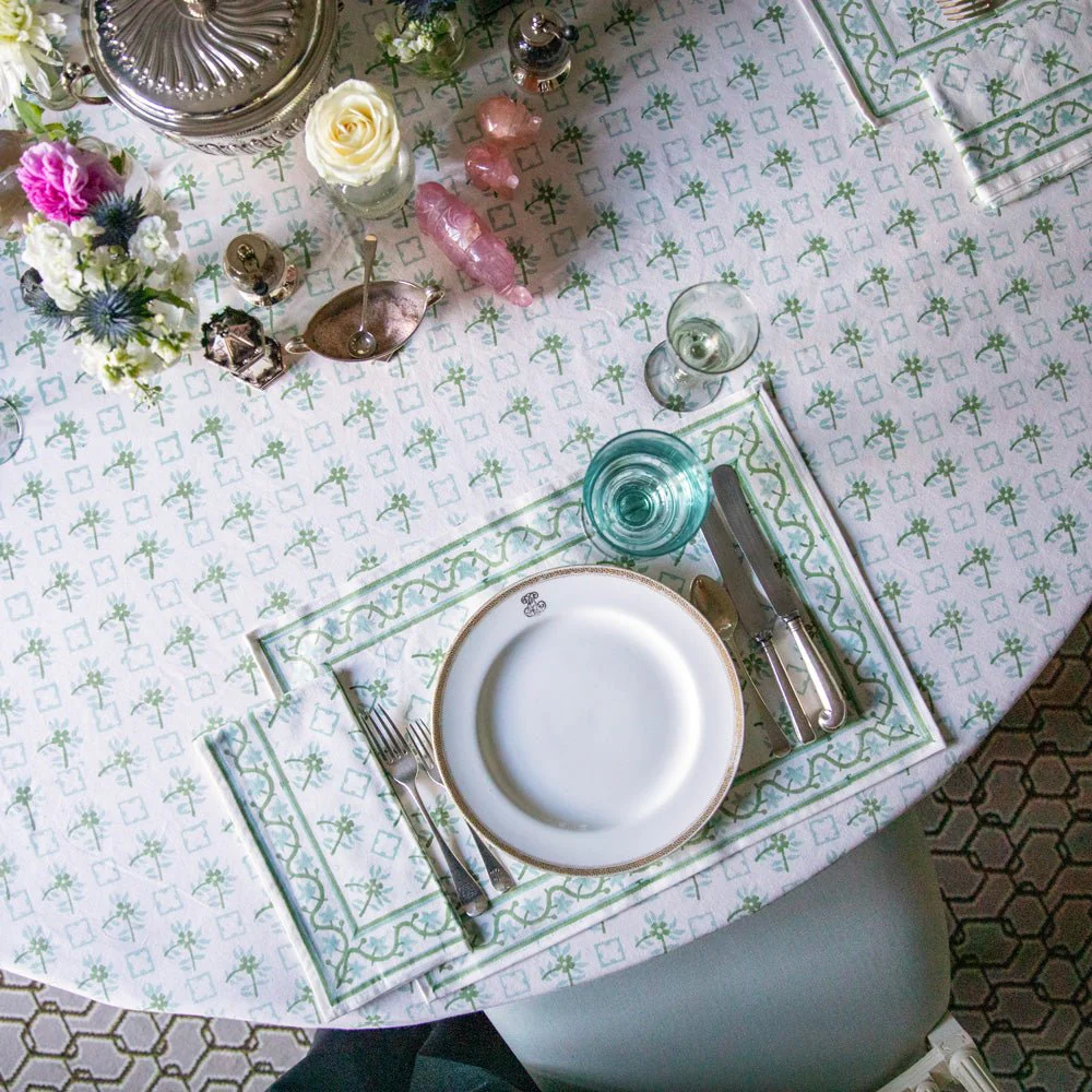A decorated table with a white patterned tablecloth, set with a white plate, silverware, a wine glass, and a water glass. The table has floral decorations including a bouquet of pink, purple, and white flowers, a white rose, and small pink and purple