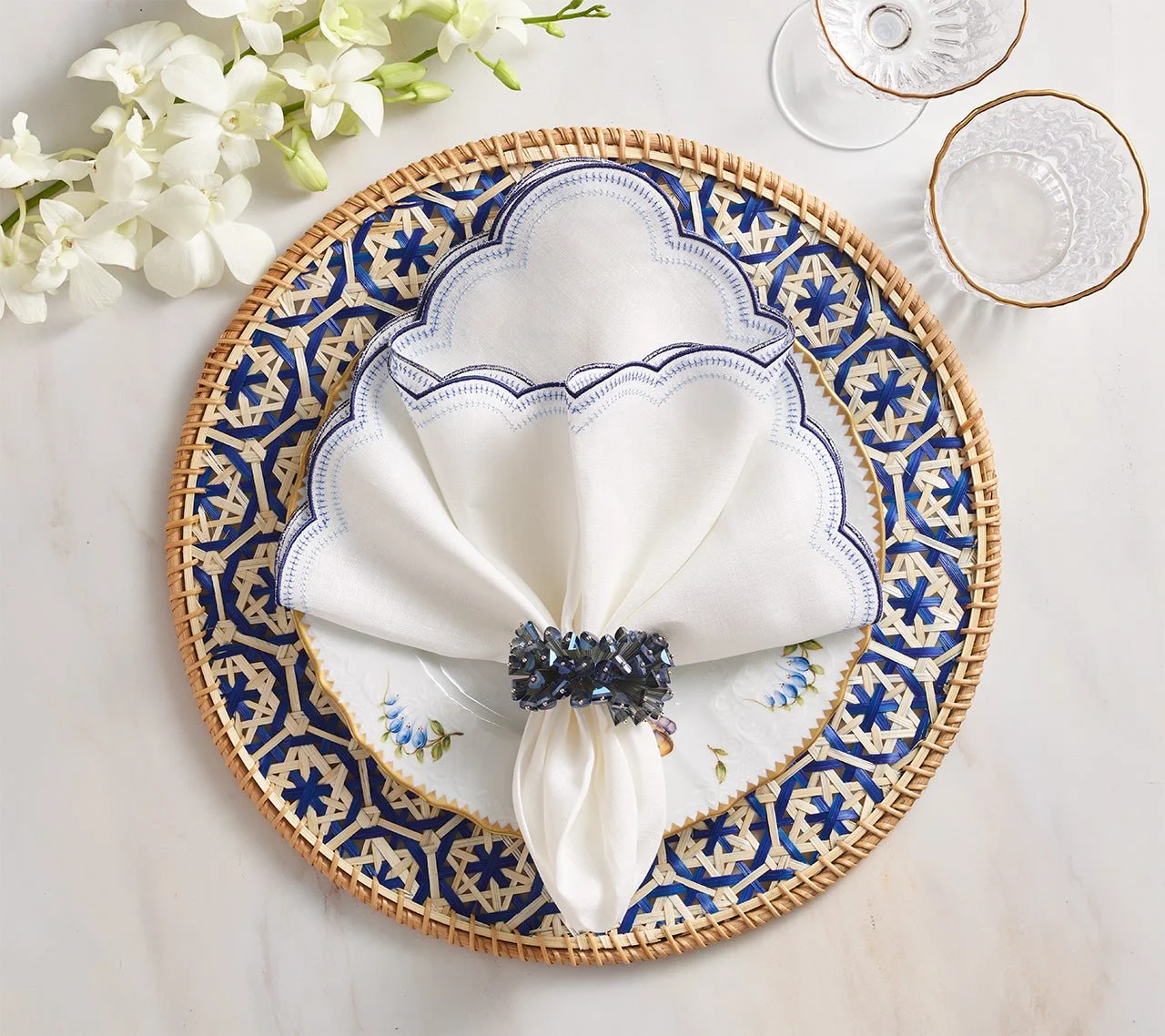A white napkin with blue embroidery is folded and held with a decorative black napkin ring, placed on a white plate with blue and yellow floral design. The plate is on a round woven placemat with a blue pattern, on a white surface with white flowers and two clear glass cups nearby.