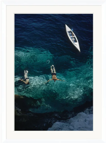 Two people swimming in clear blue water near rocks, with a white boat floating nearby.