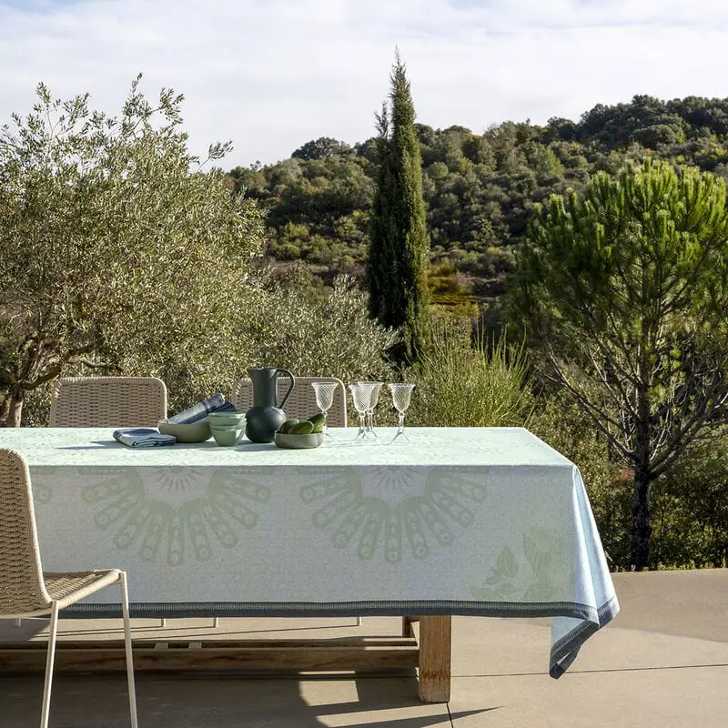 Outdoor dining table with a tablecloth, set with green bowls, a black pitcher, and three wine glasses, surrounded by outdoor chairs, overlooking a lush, hilly landscape with trees and a cloudy sky.