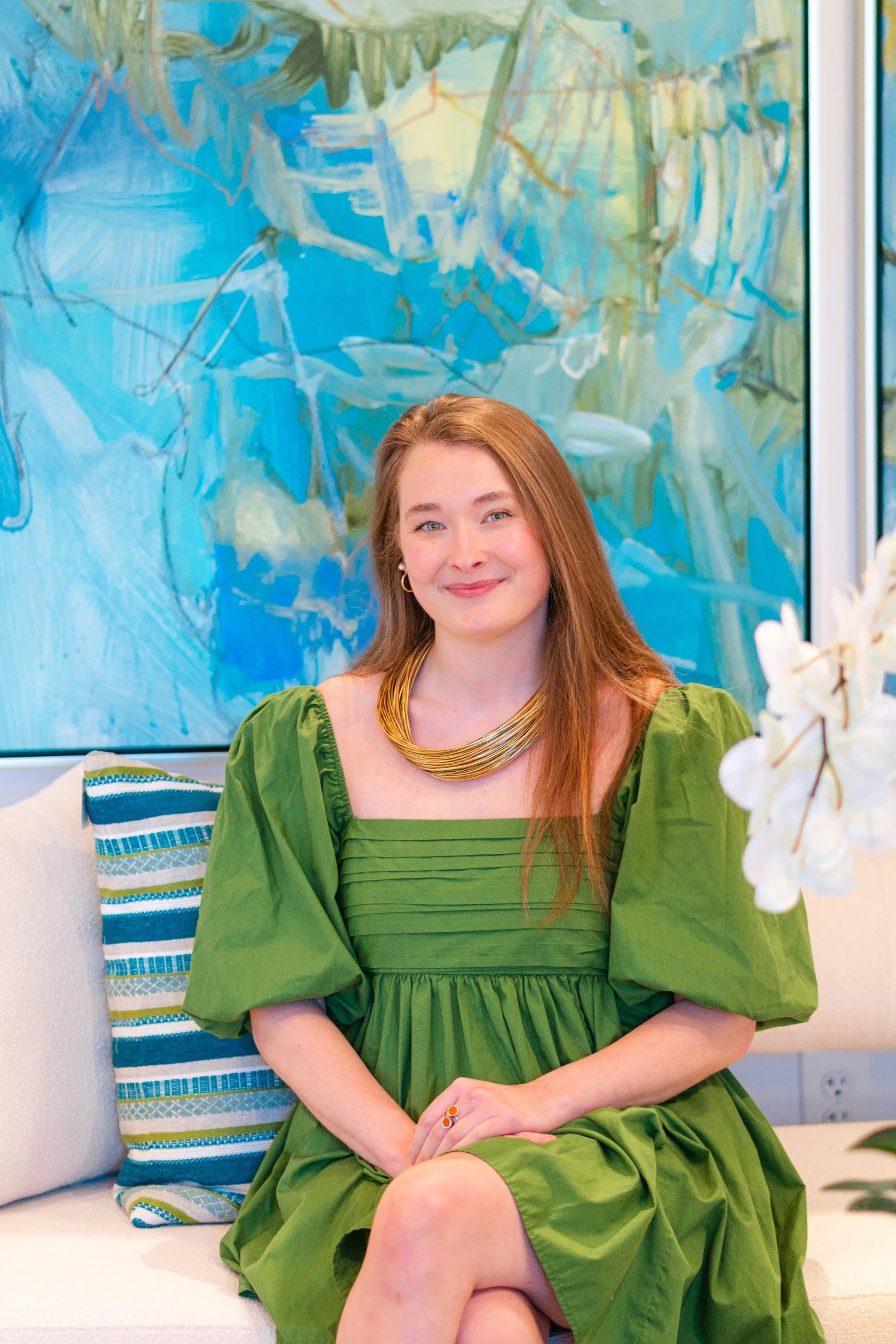 Abigail Barnard, DASH Outdoor Sales Coordinator, sitting on a modern cream sofa with a striped blue and green pillow, wearing a green puff-sleeve dress and gold necklace, in front of a vibrant abstract blue painting