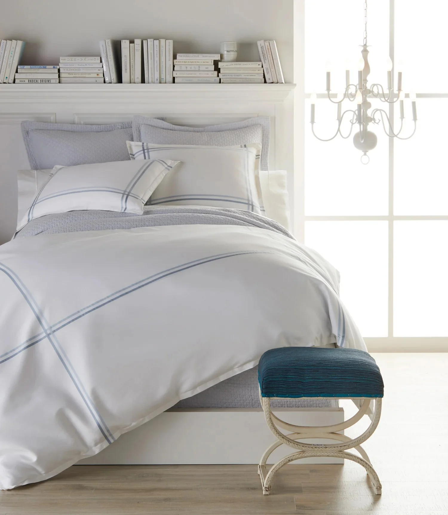 A neatly made bed with white and gray plaid bedding, set against a bright window. A small blue cushioned stool with a white curved frame is at the foot of the bed. A white chandelier hangs near the window, and a built-in shelf above the bed holds boo