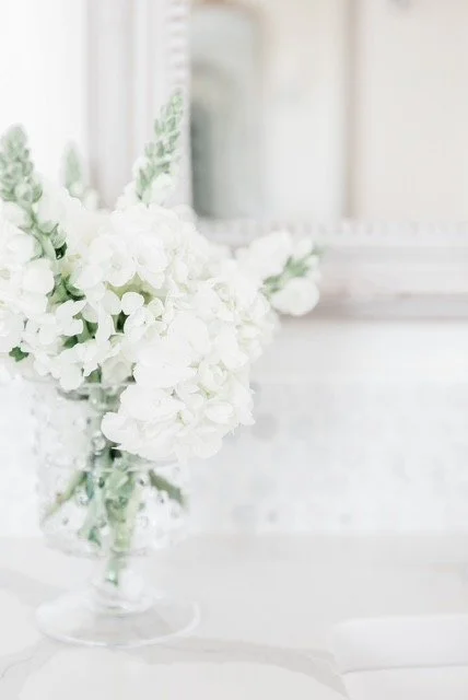 White flowers in a vase on a white table in a bright room with a mirror and decorative mirror frame in the background.