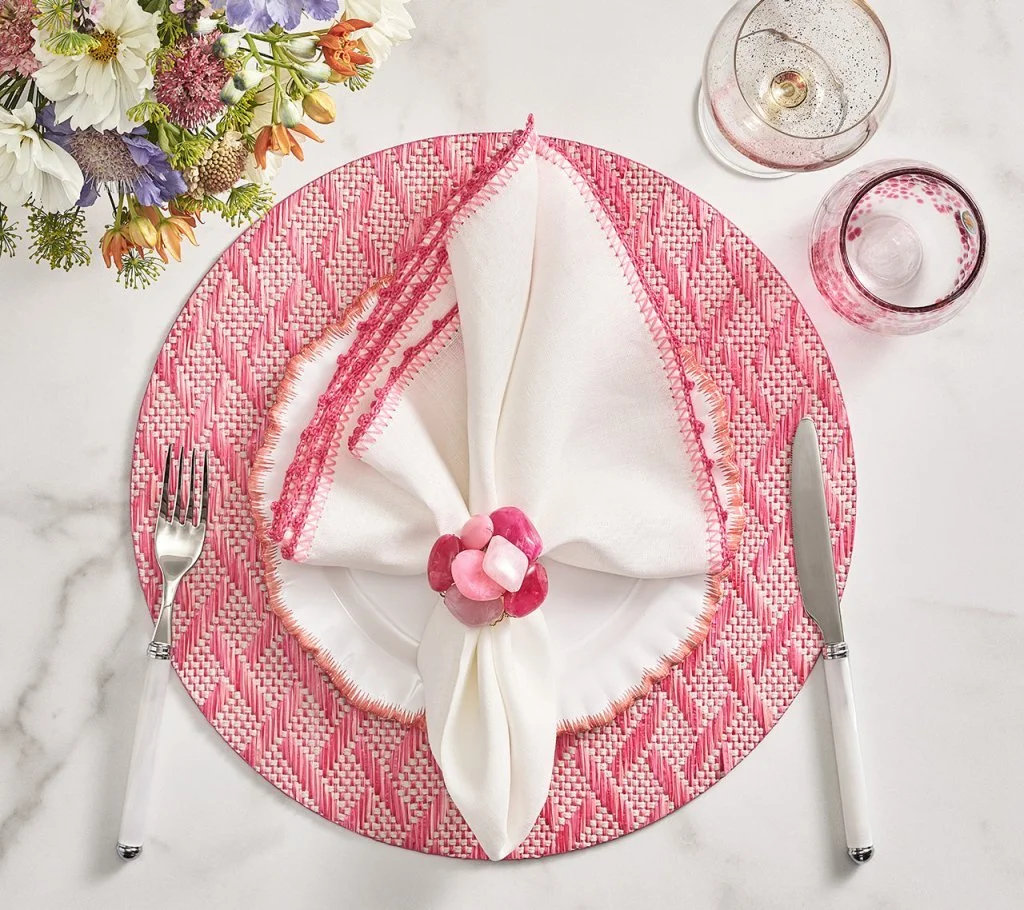 A table setting featuring a pink and white woven placemat, with a white cloth napkin folded and decorated with pink and white ornaments, on a white plate. To the left is a fork, to the right a knife, and above are two glasses, one with a bubbly drink and the other with a pink beverage. A flower arrangement with various colorful flowers is positioned at the top left corner.