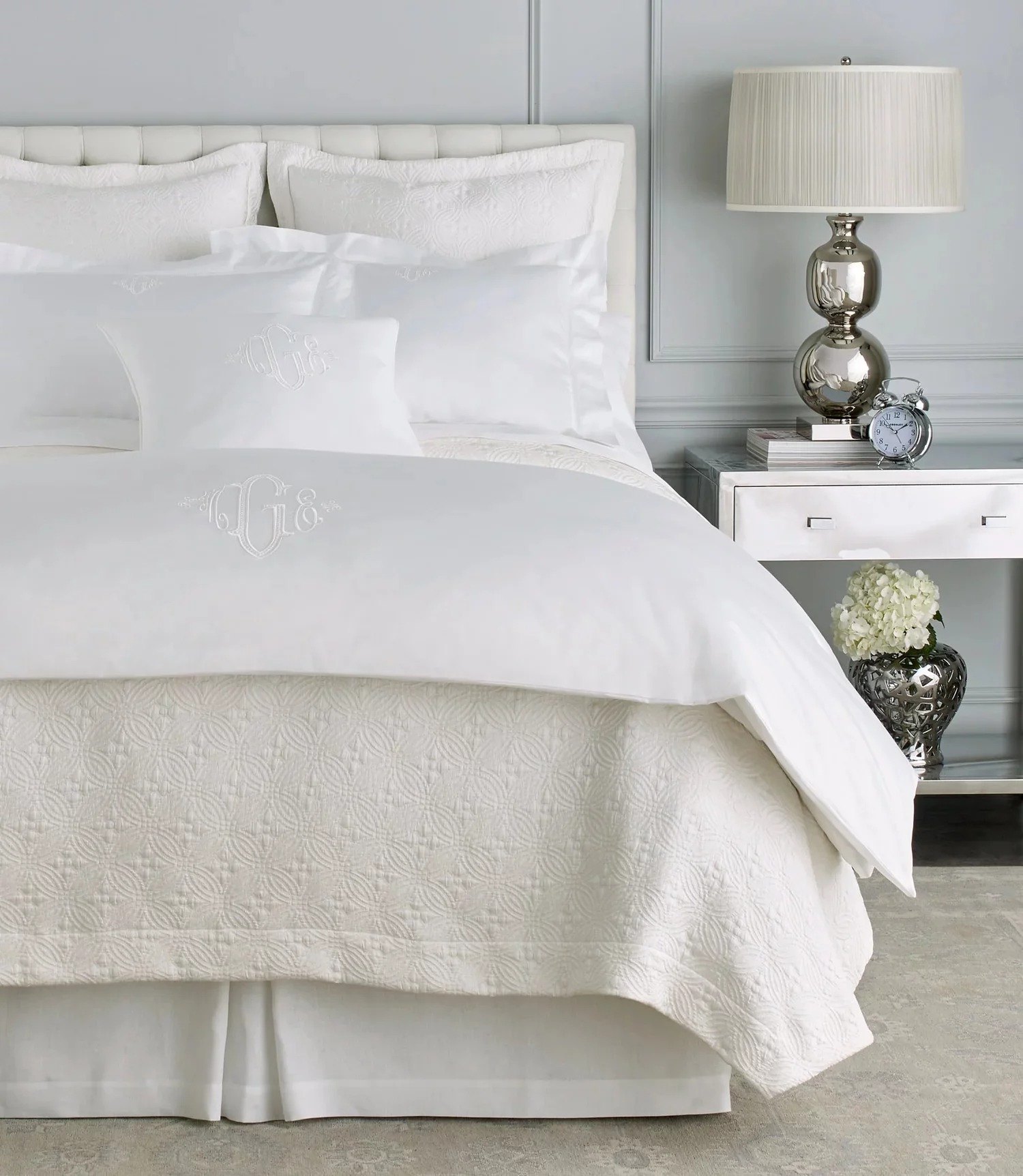 White bed with embroidered monogrammed pillows, a matching duvet, and a quilted bed skirt in a modern bedroom. Next to the bed is a white nightstand with a silver lamp, an alarm clock, and a stack of books. A silver vase with white flowers is on the