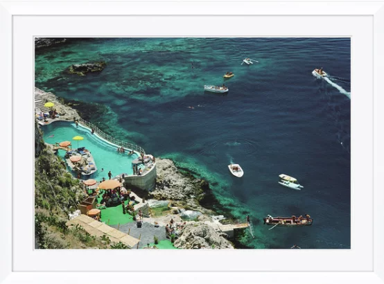 A pool near the coast with several boats on the water, some boats near the pool, and a rocky shoreline with umbrellas and people.