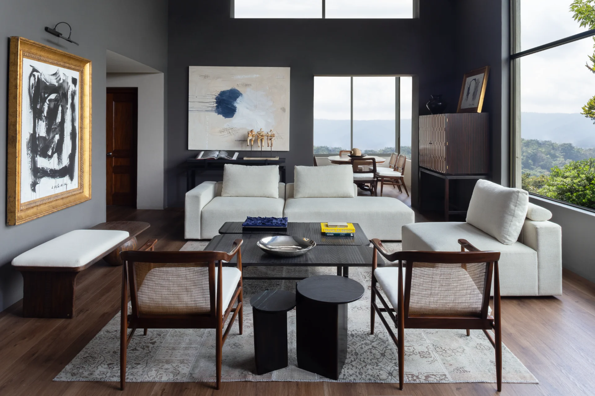 Living room with white sofa, wooden chairs, abstract paintings, and large windows showing a scenic outdoor view.