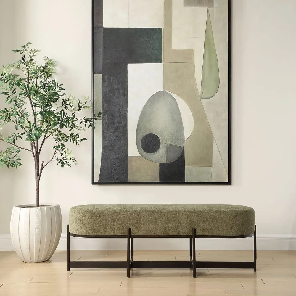 A green upholstered bench with black metal legs is placed against a white wall. To the left, there is a tall potted plant with green leaves in a white pot. Above, a large abstract painting with geometric shapes and muted colors hangs on the wall.