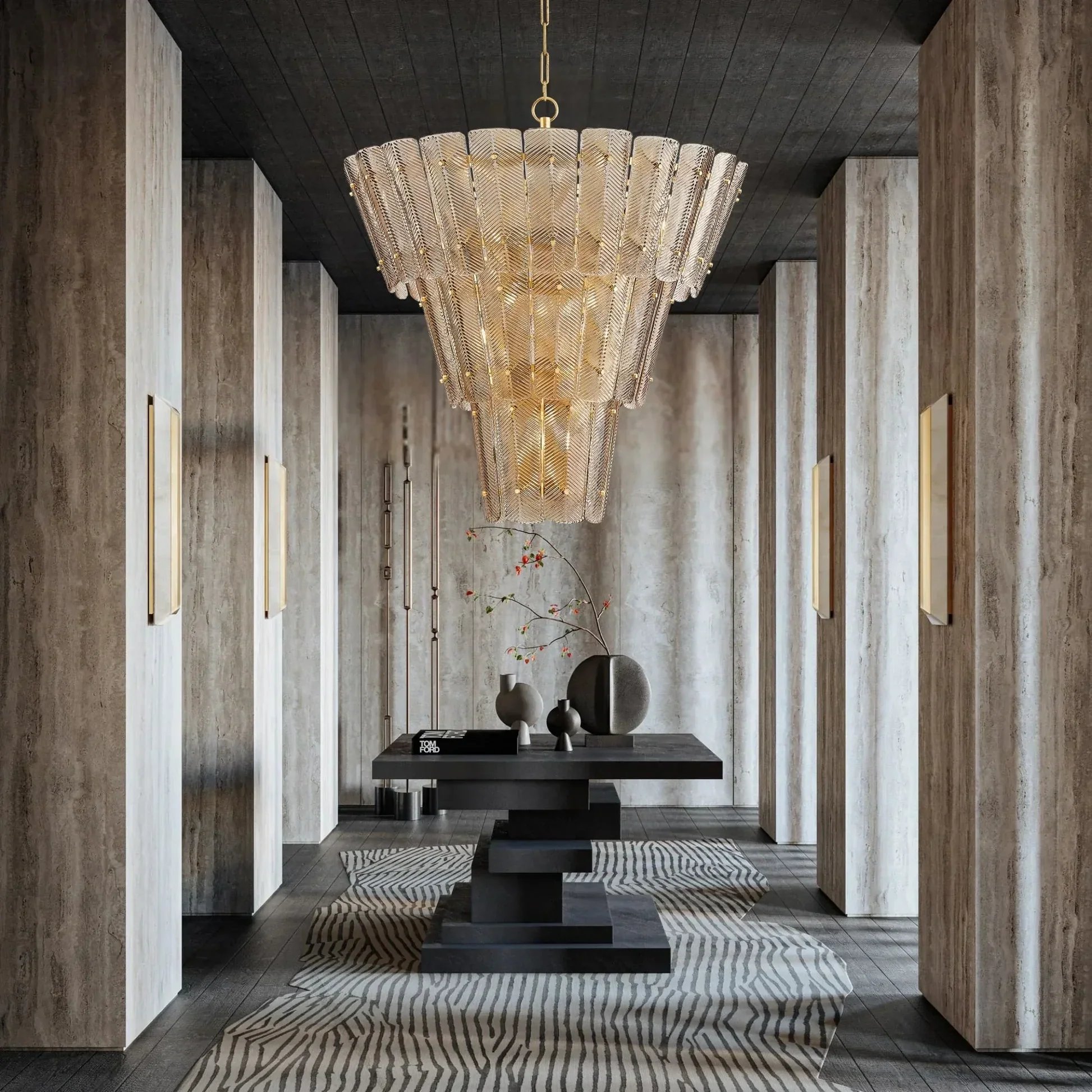 A large gold chandelier with a feather design hangs from a black ceiling in a modern interior space. The room features beige stone walls with vertical gold accents, dark wood flooring, and a black table decorated with various vases and a sign that reads 'Tom Ford.' There is a plant with red flowers and a patterned rug on the floor.