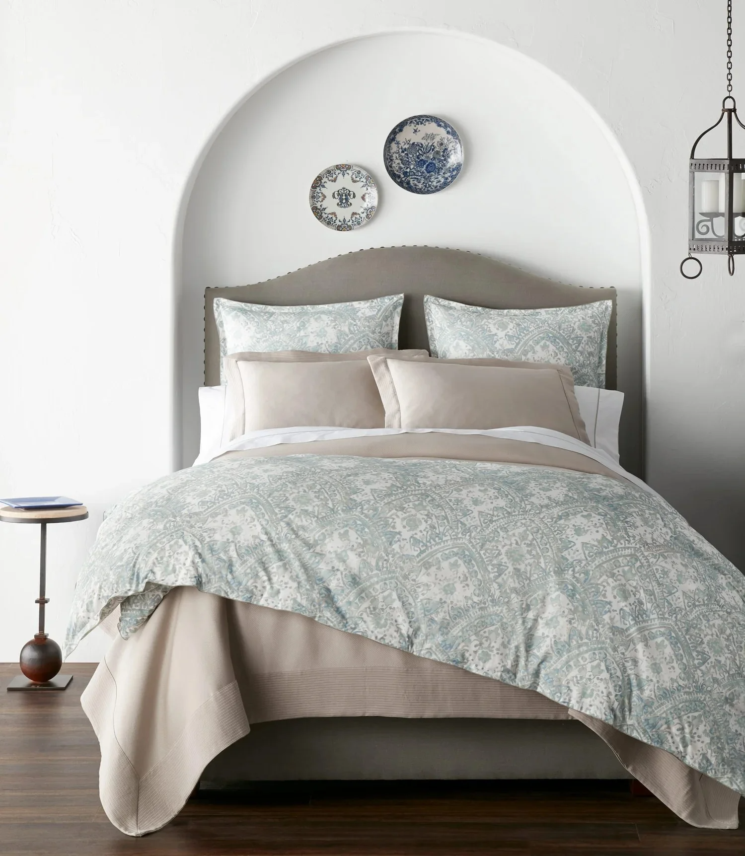 A neatly made bed with patterned bedding and multiple pillows, framed by a white arched wall with decorative plates hanging above, a side table with a book on it to the left, and a lantern on the wall to the right.