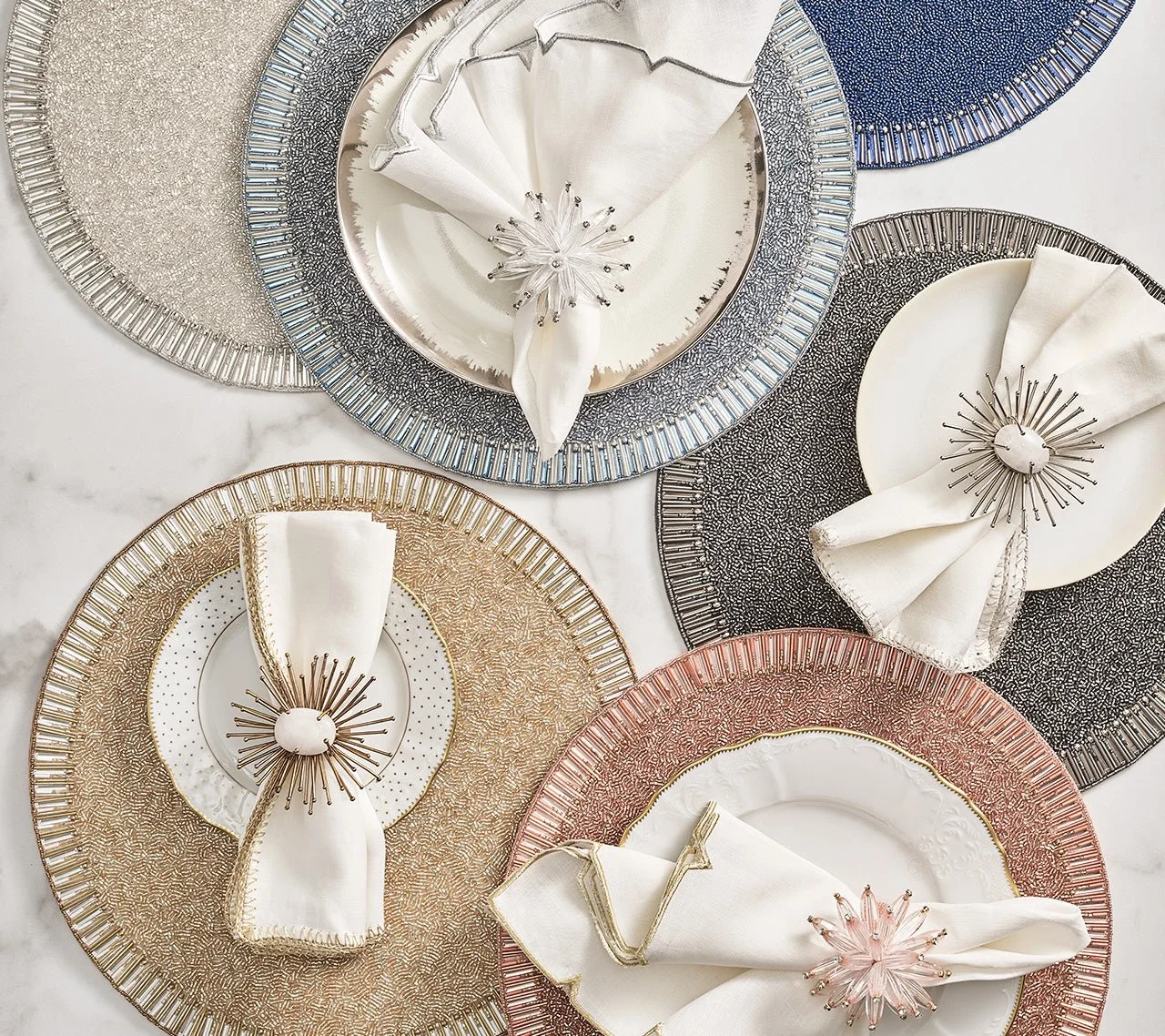 Colorful decorative plates with detailed patterns, each paired with white napkins secured by metallic napkin rings, arranged on a white marble surface.