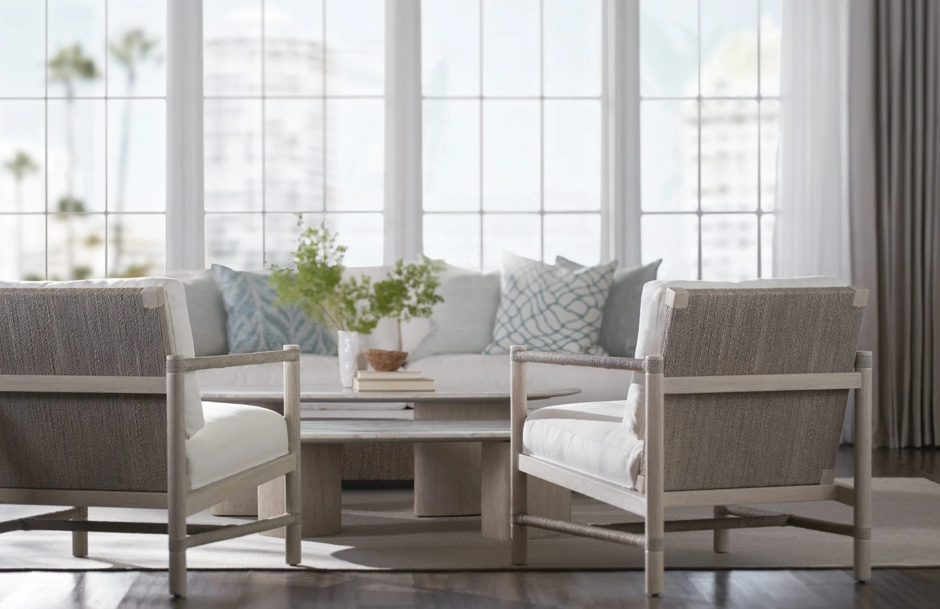 Living room with a sofa, armchairs, and a coffee table in front of large windows with sheer curtains, decorated with pillows and a potted plant.
