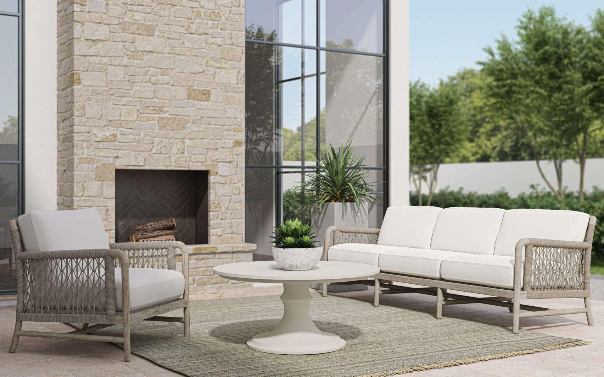 Outdoor patio with white seating, a round white table with plants, a stone fireplace, large windows, and green trees outside.