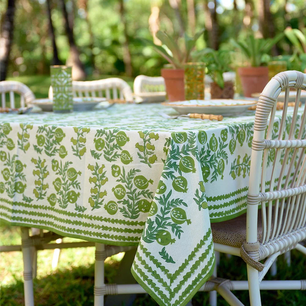 Outdoor table with a green and white patterned tablecloth, set with plates and glasses, surrounded by white wicker chairs, with potted plants and trees in the background.